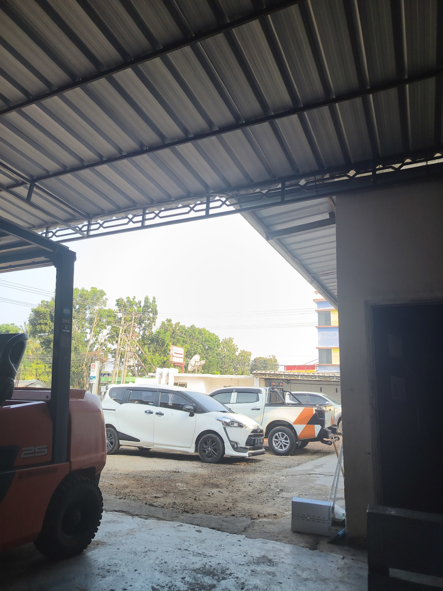 The image captures a section of an outdoor covered area, possibly a workshop or storage space, with a corrugated metal roof supported by metal beams. In the foreground, the side of an orange forklift with the number "25" is visible on its left side. Beyond the forklift, parked on a dirt and concrete surface, are two white vehicles: a sedan and a pickup truck. The sedan is white and appears to be a multi-purpose vehicle, while the pickup truck has a distinctive orange and white graphic design on its rear. In the background, lush green trees and utility poles are visible under a bright, overcast sky. A multi-story building with yellow and blue accents can be seen further in the distance. The scene suggests an industrial or commercial setting in Pangkalpinang, Indonesia, during daylight hours.