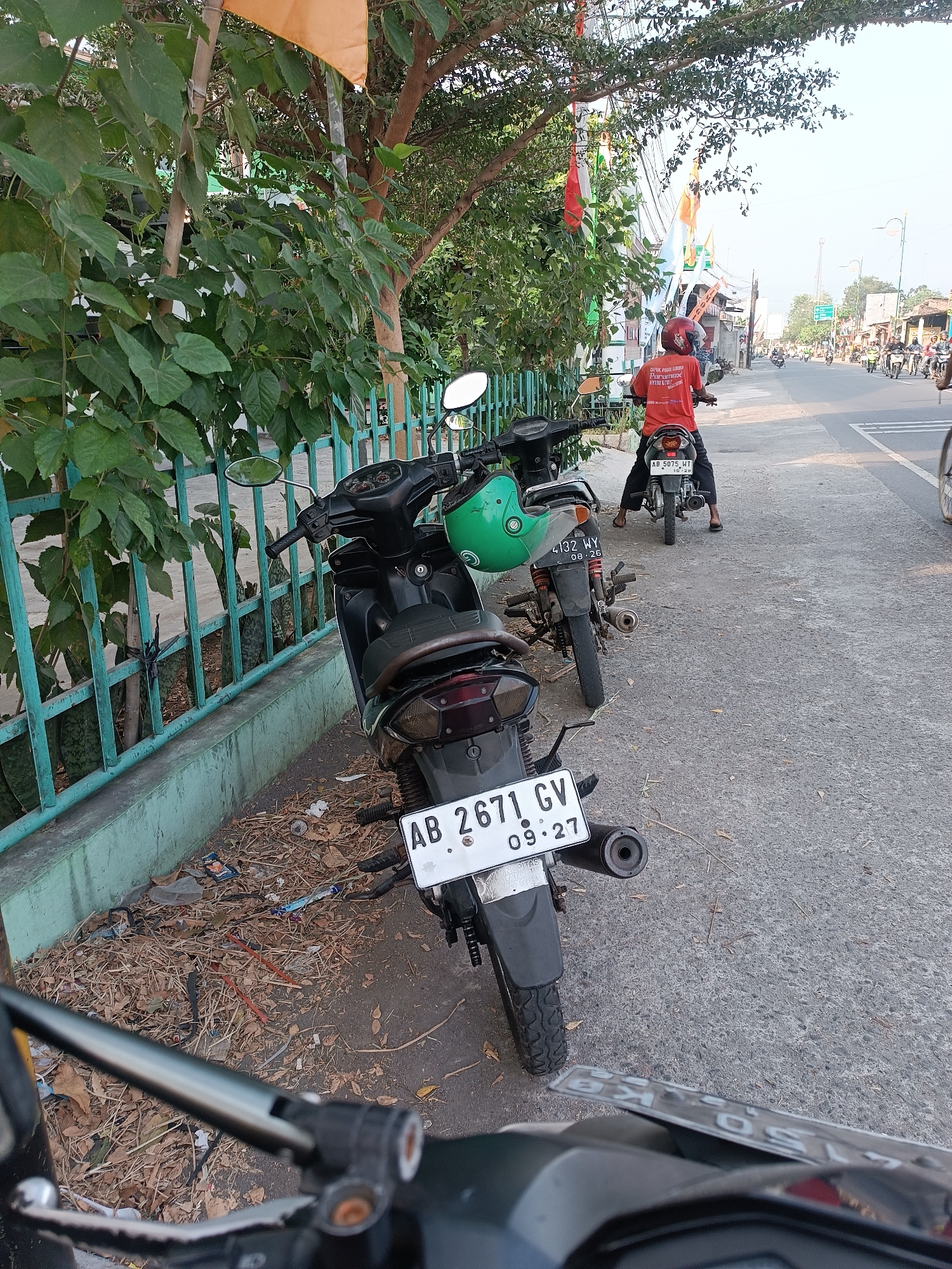 In Imogiri, Indonesia, along a sun-drenched roadside, a scene unfolds with several parked motorcycles. The foreground is dominated by the rear of a black motorcycle, its license plate reading "AB 2671 GV 09.27". To its left, another motorcycle is partially visible, with a green helmet resting on its handlebars. Further down the street, a person wearing a red t-shirt and helmet is seated on a motorcycle, facing away from the camera. The street is lined with green metal fencing and lush green foliage. In the background, more motorcycles and people can be seen, suggesting a bustling street environment. The ground is covered with dry leaves and scattered debris, indicating a potentially dry season. The overall impression is of a typical day in a vibrant Indonesian town, with the ubiquitous presence of motorcycles defining the landscape.