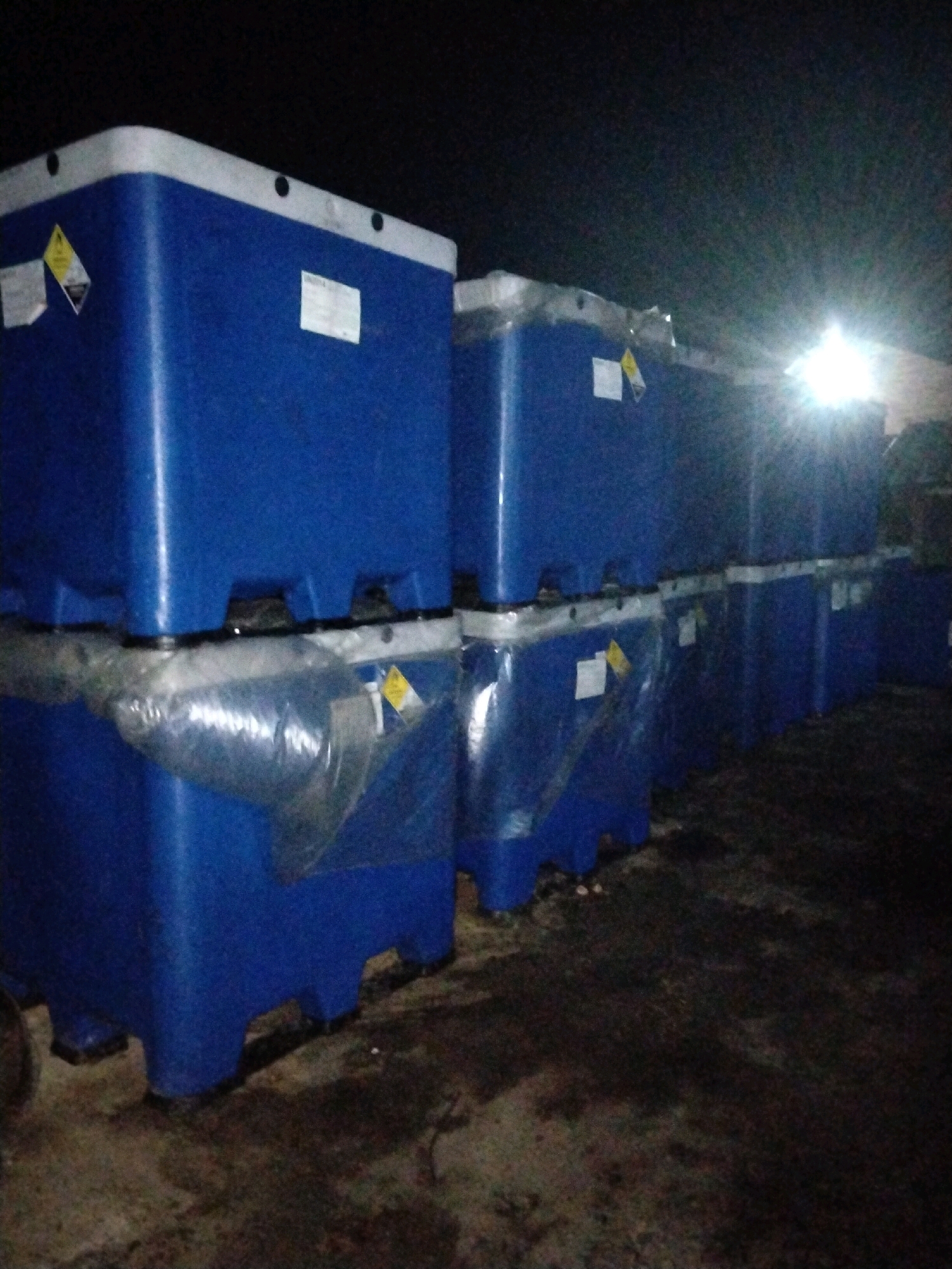 A pile of blue industrial plastic containers is stacked in an outdoor area. The containers are approximately 1 cubic meter in volume, with a white top rim and a dark blue body. They are covered in clear plastic wrap, and some have yellow hazard symbols and white labels attached. The ground appears to be concrete or asphalt, and it is dark, suggesting the image was taken at night or in low light conditions. There is a bright light source in the background, possibly a streetlamp or artificial lighting. The scene is located in the city of Moro, Indonesia.