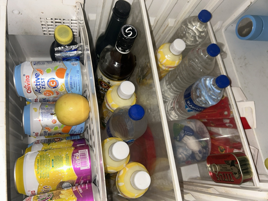 An open refrigerator interior in Egbe, Nigeria, displays three visible shelves containing various beverages and food items.
The top shelf contains several upright cans. On the left, multiple yellow and purple cans labeled "Oluglic Sparkling Pineapple Coconut" are visible. To their right, several light blue and white cans of "Chivita Active Zest Mixed Fruit Sparkling" are present. A single yellow-skinned fruit, possibly a passion fruit, rests among these cans. On the far right, a yellow bottle cap lies near a crumpled silver wrapper and a dark, indistinct object. The shelf surface appears slightly stained.
The middle shelf holds multiple items horizontally. On the left, three plastic bottles containing a yellow liquid with white caps are arranged. Adjacent to these is one clear plastic bottle with a blue cap, containing clear liquid. To the right, two dark glass bottles are present: one brown bottle with a black and silver cap (marked with an 'S'), and one black bottle with a black cap. The shelf surface shows some dirt and residue.
The bottom shelf contains a red rectangular tin labeled "TITUS SARDINES" on its left side. In the center, a clear plastic container holds various small, crumpled wrappers, with a red object behind it. To the right, multiple plastic bottles, primarily "AQUAVIE" brand, contain clear liquid and have blue caps; one bottle with yellow liquid and a white cap is also present. On the far right, a light blue cylindrical object, possibly a reusable cup or thermos, stands upright. The bottom shelf and floor of the refrigerator appear significantly dirty with visible grime. No people are present in the scene.