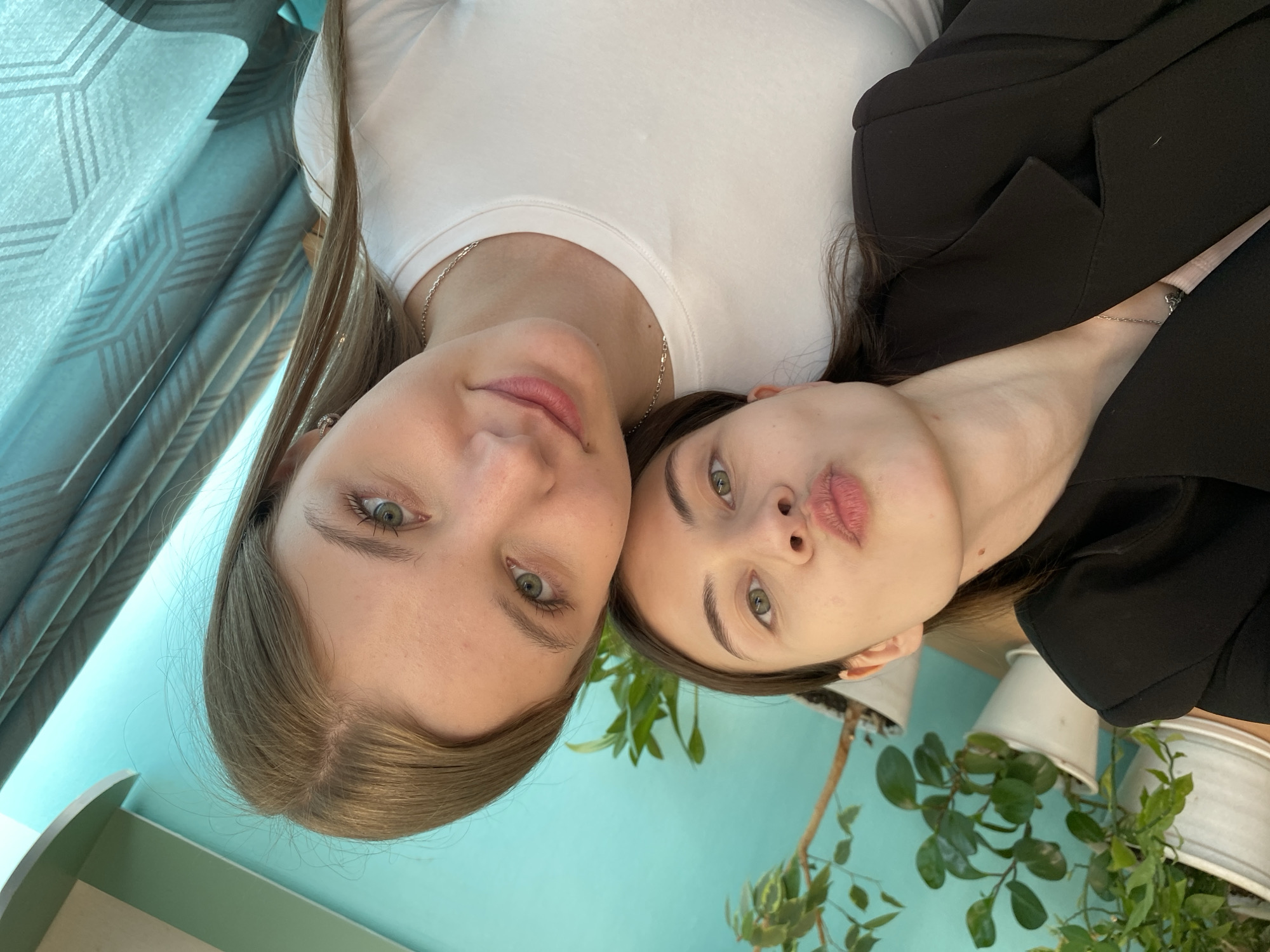 Two women are close together, appearing to be posing for a selfie. The woman on the left has light brown hair, fair skin, and green eyes. She wears a white t-shirt and a silver necklace. The woman on the right has dark brown hair, fair skin, and green eyes. She wears a black blazer and a silver necklace. The background features a teal wall, a curtain, and plants in white pots. While the location is given as Votkinsk, Russia, there are no specific visual markers in the image that confirm this. The image appears to be taken indoors. The women's heads are close together, with their faces near each other.
