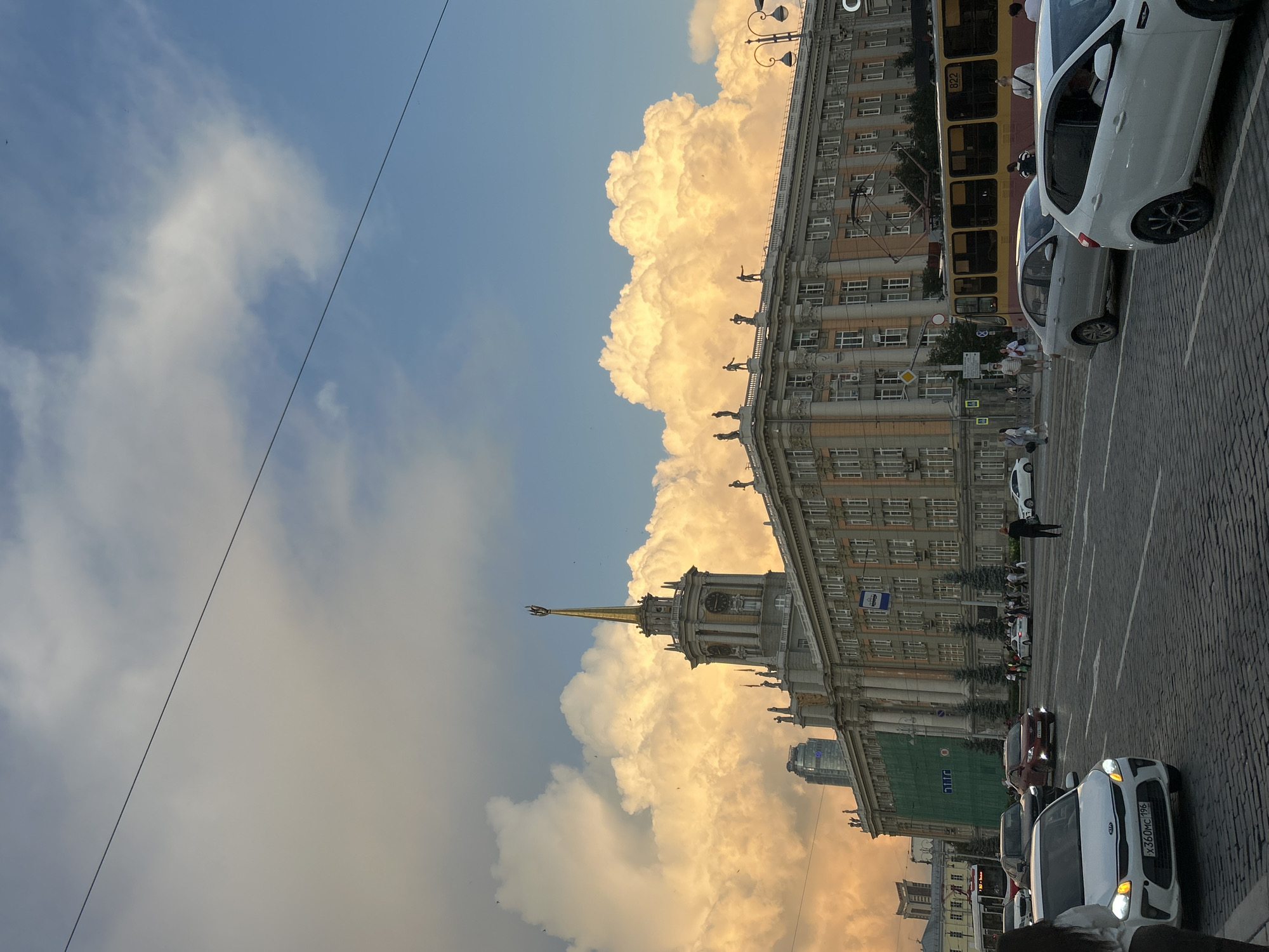 The image depicts a street scene in Yekaterinburg, Russia, likely near the Yekaterinburg City Administration building, also known as the "House of Soviets." A large, ornate building with a central tower topped by a golden spire dominates the background. The building features classical architectural elements, including statues of figures adorning its roofline and columns. A bright, dramatic sky with large, illuminated clouds, possibly a sunset or storm light, is visible above the building.
In the foreground, a cobblestone street is filled with traffic. Several cars, including a white Kia with a license plate starting with "X360MC," are in motion. A yellow and red tram with the number "822" is also present, traveling along its tracks. Pedestrians are visible on the sidewalks and crossing the street. The overall atmosphere suggests a busy urban environment during a visually striking weather event.