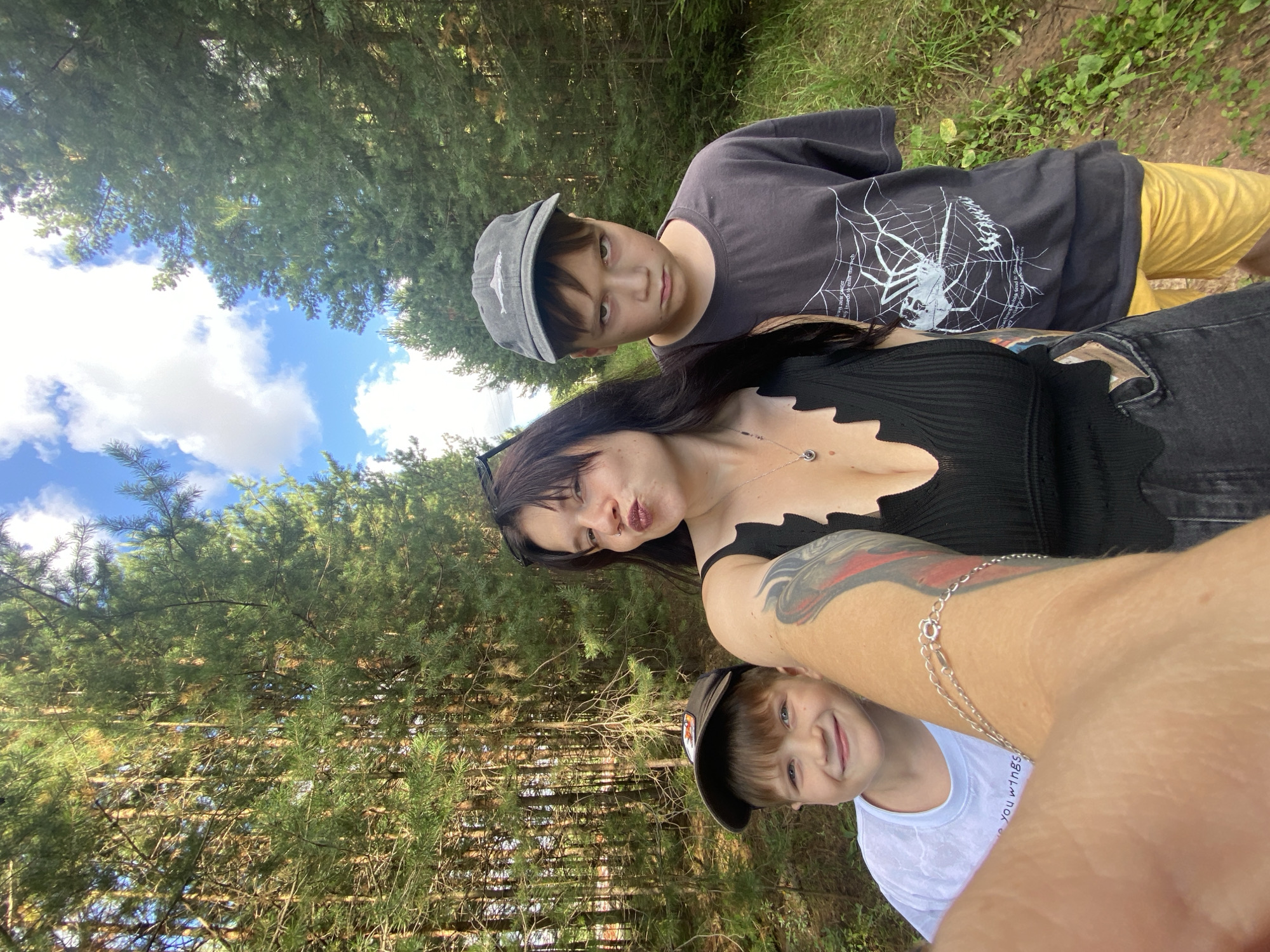 A young woman with dark hair is taking a selfie with two young boys in a forest. The woman is wearing a black tank top and jeans. The boy on the left is wearing a white t-shirt and a baseball cap. The boy on the right is wearing a dark t-shirt with a spiderweb graphic and yellow shorts, and a baseball cap. The trees are tall and green, and the sky is blue with white clouds. The ground is covered with grass and dirt. The image appears to have been taken in the city of Fokino, Russia.