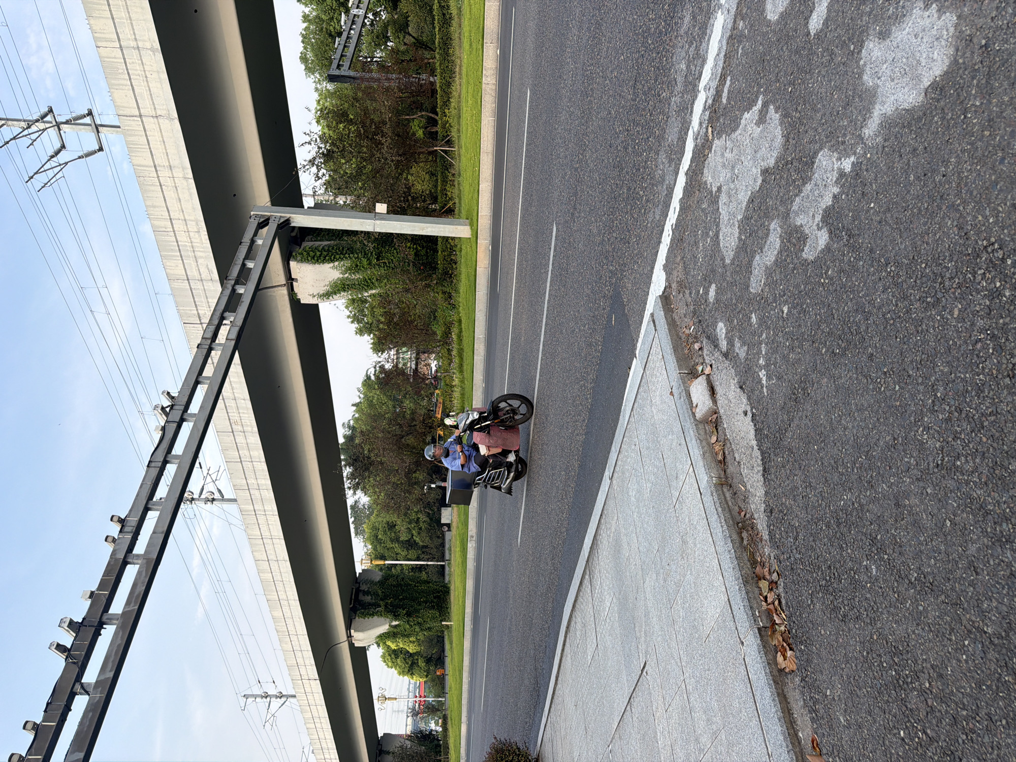 An elderly person wearing a blue long-sleeved shirt and a light-colored helmet is riding a black electric scooter on a paved road. The scooter has a large black cargo box mounted on the rear. The rider is looking forward and appears to be traveling on the right side of the road. The road is multi-lane with white lane markings. To the left of the road is a sidewalk made of gray tiles, bordered by a concrete curb. Fallen brown leaves are scattered along the curb. In the background, a large concrete overpass stretches across the scene, supported by concrete pillars. Above the overpass, power lines and support structures are visible against a pale blue sky. Green trees and bushes line the area behind the road. The overall setting suggests an urban environment, likely within Jiangjin, China, given the context.