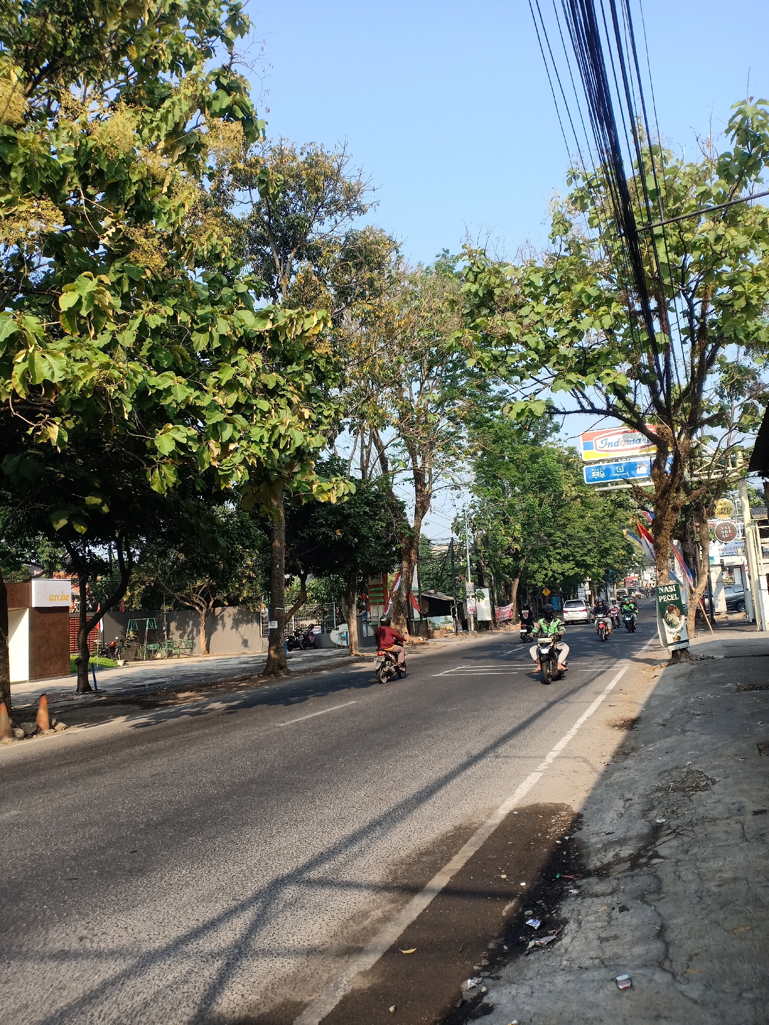 The image depicts a street scene in Semarang, Indonesia. The view is of a road with traffic, primarily motorcycles. Tall trees with dense green foliage line the left side of the street, casting shadows across the asphalt. On the right side, there are buildings and a prominent Indomaret convenience store sign. A few people are visible riding motorcycles, with one individual on the far left of the frame and several others in the middle and right portions of the road. The sky is clear and blue, indicating daytime. The overall impression is of a moderately busy urban street.