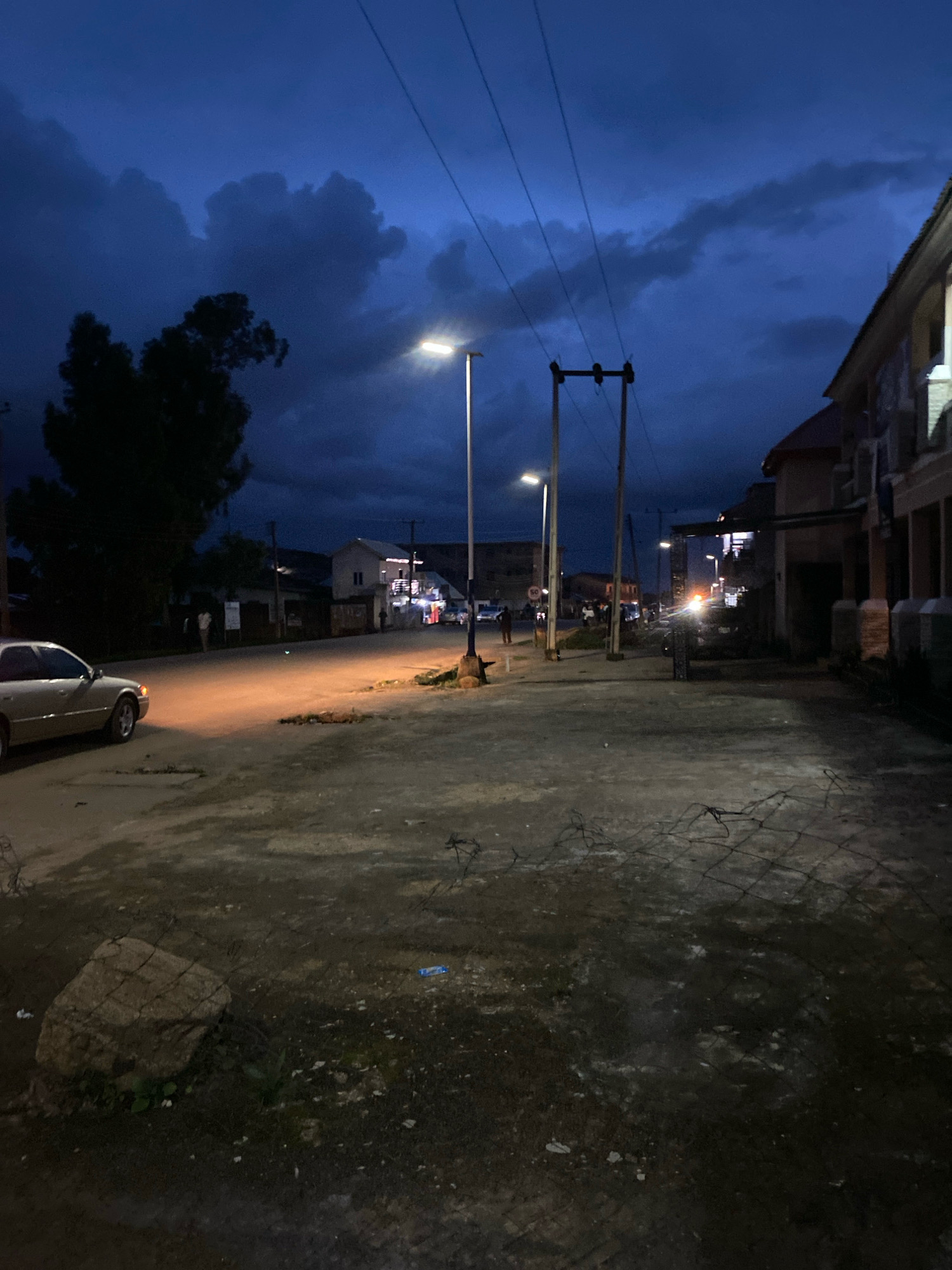 This is a nighttime image of a street scene in Jos, Nigeria. The sky is dark blue with dramatic clouds, illuminated by streetlights. A light-colored sedan is parked on the left side of the unpaved road, its headlights casting a warm glow. Several people are visible as indistinct figures on the sidewalk and in the road, some near buildings. The buildings on the right appear to be commercial or residential structures with illuminated windows. Utility poles and overhead wires traverse the scene. The foreground shows a rough, uneven surface with a large rock and some scattered debris, including what appears to be wire fencing. The overall impression is of a quiet street at dusk or early night.