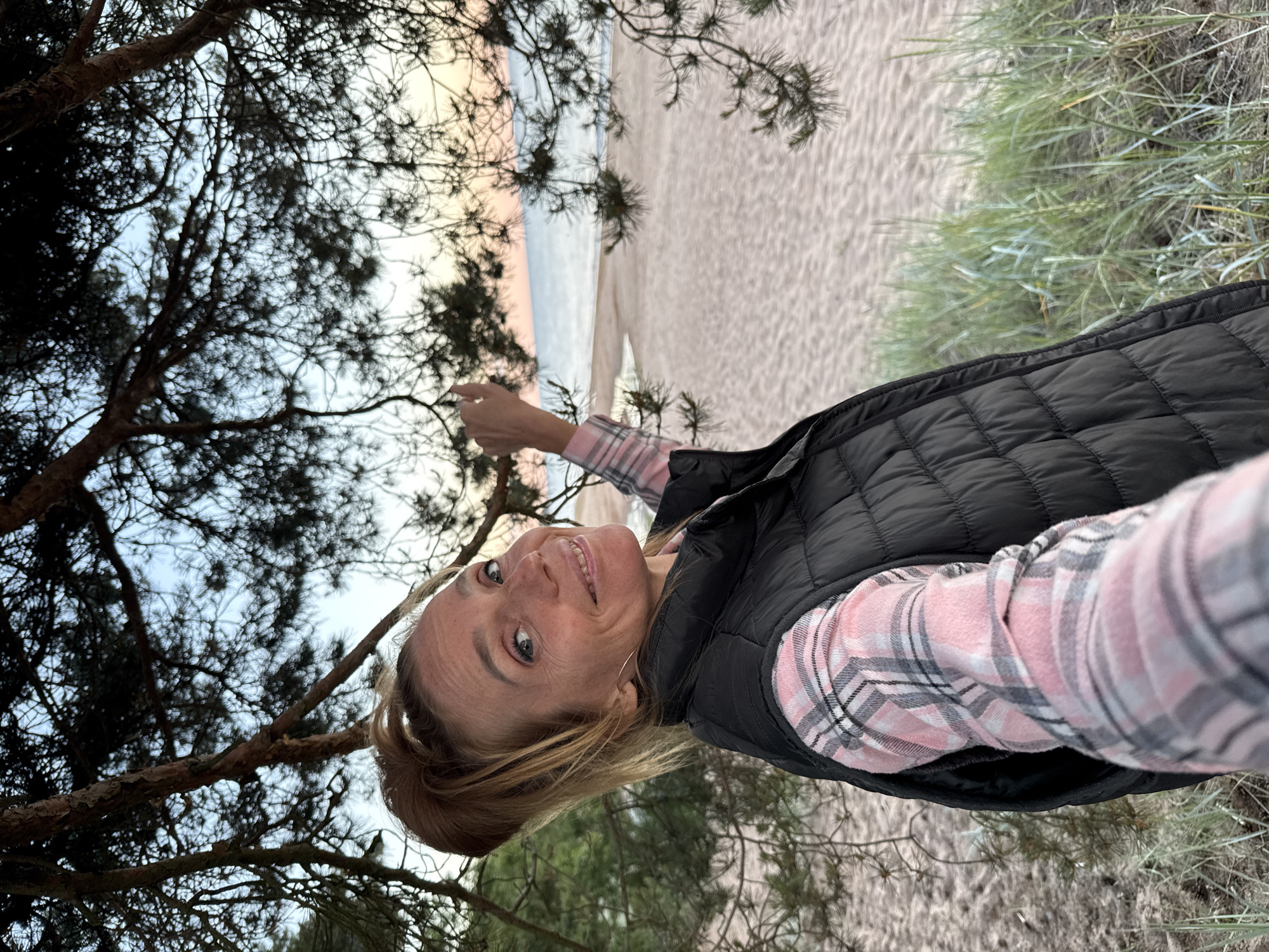A blonde woman stands on a sandy beach, partially obscured by pine trees. She is wearing a pink and gray plaid shirt and a black puffer vest. Her hair is pulled back in a ponytail. She is smiling and looking at the camera, with her right arm extended. In the background, there is a calm sea and a soft sunset sky. The location is the beach in Zelenaya Roshcha, Russia.