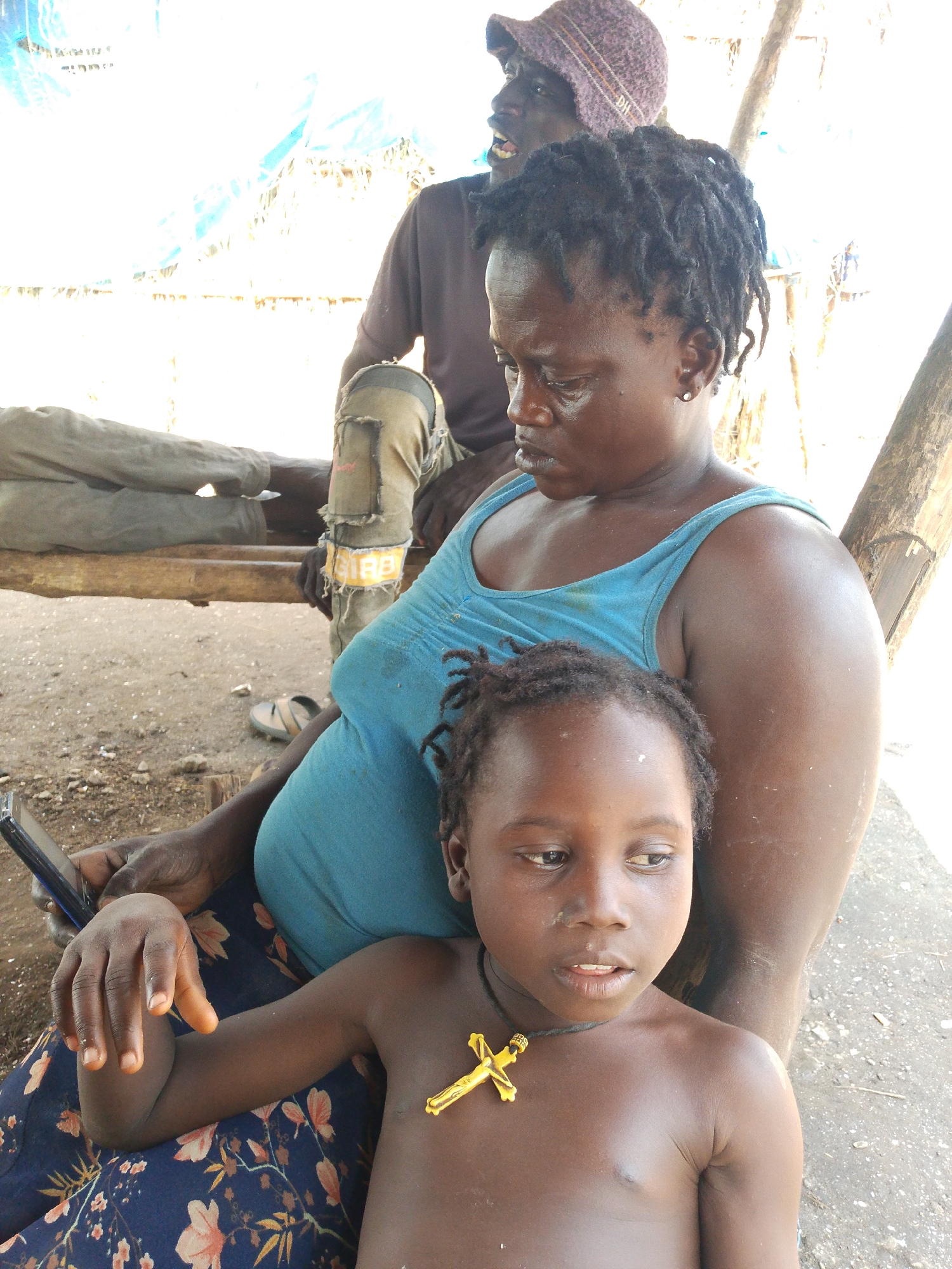 In Otulu, Nigeria, a group of individuals are seen gathered, possibly in a rural setting. A woman in a blue tank top is holding a young child who is shirtless and wearing a gold cross necklace. The woman has dark hair styled in dreadlocks and appears to be looking down. Beside her, a person wearing a purple knitted hat and a brown shirt is partially visible, with their mouth open. Another individual is seated to the left, wearing worn brown pants. The ground appears to be dusty and uneven, with some debris scattered around. A wooden beam is visible in the background, possibly part of a shelter or structure. The overall lighting suggests an outdoor daytime scene.