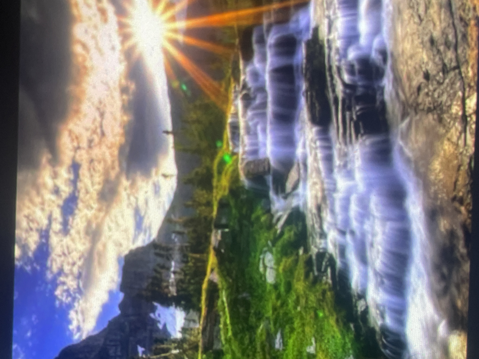 The image displays a vibrant natural landscape scene. Rugged mountains with patches of snow are visible on the left side, beneath a partially cloudy sky. The middle ground is covered in lush green vegetation, including grass and scattered coniferous trees. A multi-tiered waterfall cascades over dark rock formations in the foreground, exhibiting a long-exposure effect that renders the flowing water smooth and light-colored. A prominent sun flare with distinct rays radiates from the upper right portion of the frame, casting a warm light across the scene. No individuals or man-made structures are discernible within the image.