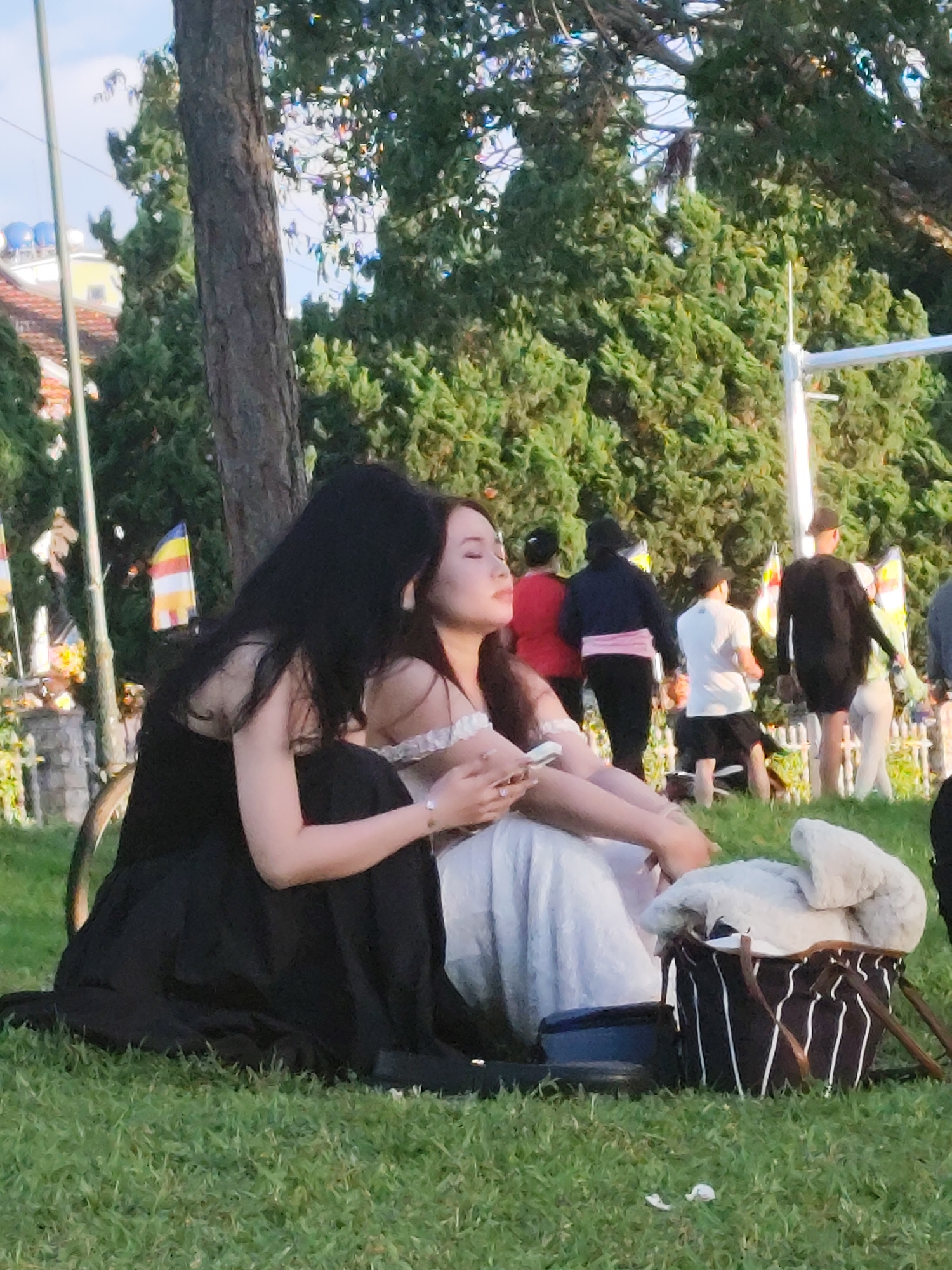 In Da Lat, Vietnam, two young women are seen sitting on a grassy lawn next to a large tree. The woman on the left, with long black hair, is wearing a black dress and is leaning towards the woman on the right. The woman on the right, with lighter hair, is wearing a white dress and is holding a smartphone. The background shows more greenery, buildings, and other people in the distance, suggesting a public park or gathering place. The atmosphere appears relaxed and casual.