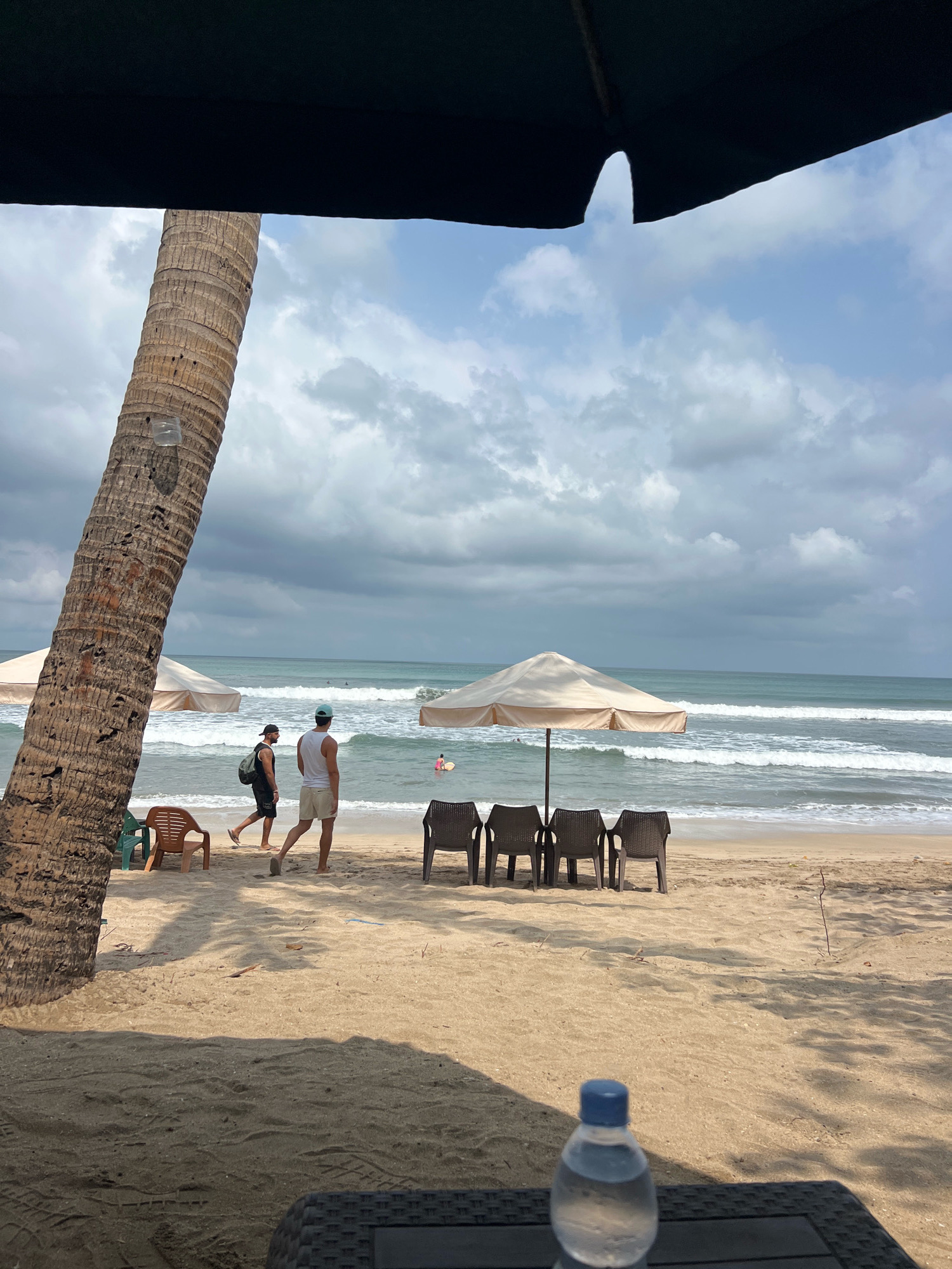 The image depicts a serene beach scene on a cloudy day in Kuta, Indonesia. In the foreground, a partially visible black umbrella casts a shadow over a plastic table holding a water bottle with a blue cap. To the left, a large, textured palm tree trunk dominates the frame, with a small plastic container attached to it. Two men walk along the sandy shore, one wearing a black tank top and shorts, and the other in a white tank top and light-colored shorts, carrying a backpack. Further down the beach, several empty lounge chairs are arranged near a beige umbrella. The ocean stretches out beyond the beach, with gentle waves rolling in under a sky filled with dramatic, gray clouds. In the distance, a few people are visible in the water, enjoying the sea. The overall atmosphere is one of relaxed tranquility.