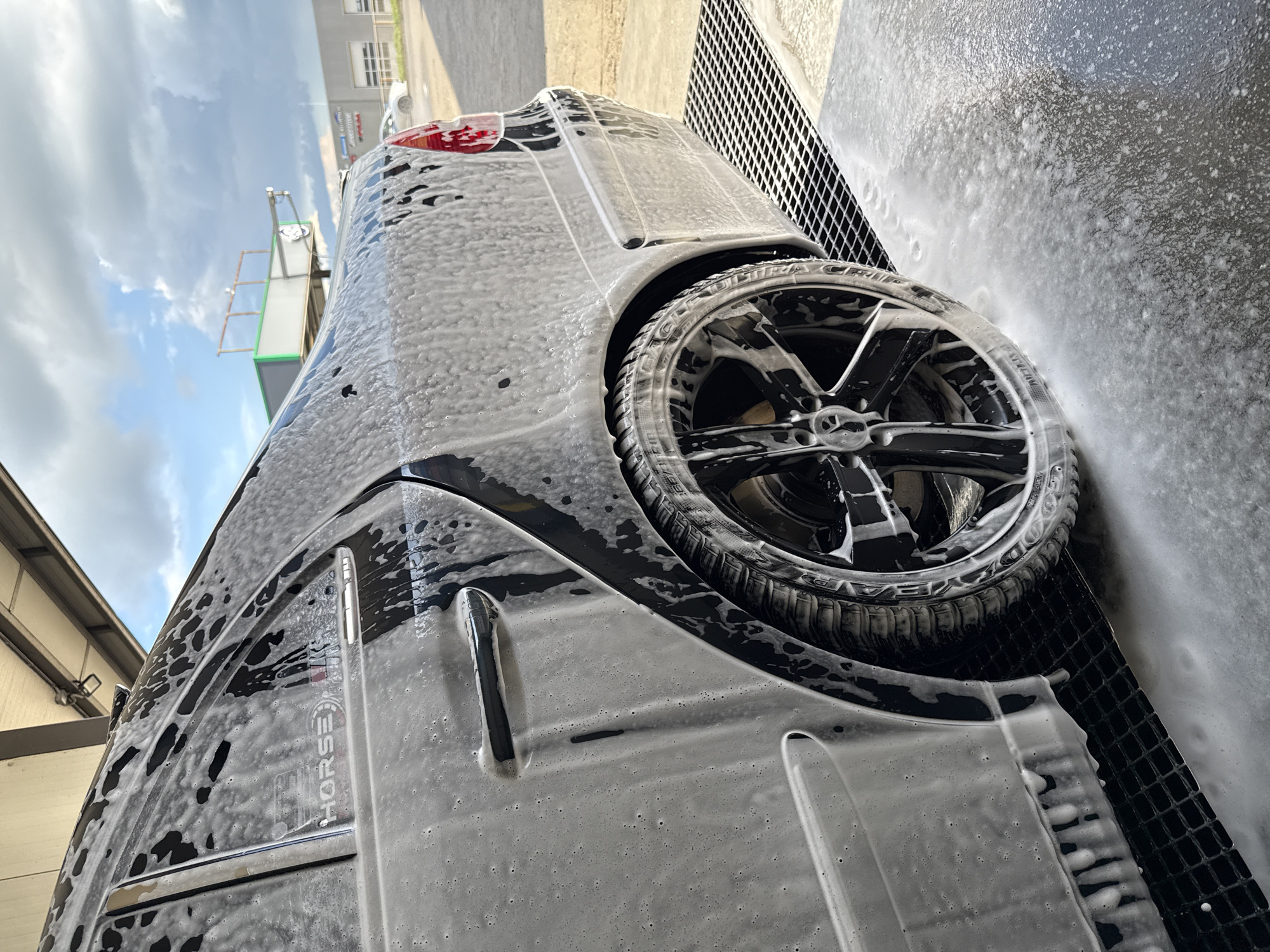 A black car is being washed with soap, obscuring details like the model. The vehicle's wheel and rear panel are visible, the latter bearing the word "HORSE" in uppercase letters. The location is Stara Zagora, Bulgaria, indicated by text on the vehicle. The car is positioned near a grate, likely part of a car wash facility. The surrounding environment includes buildings and cloudy skies.