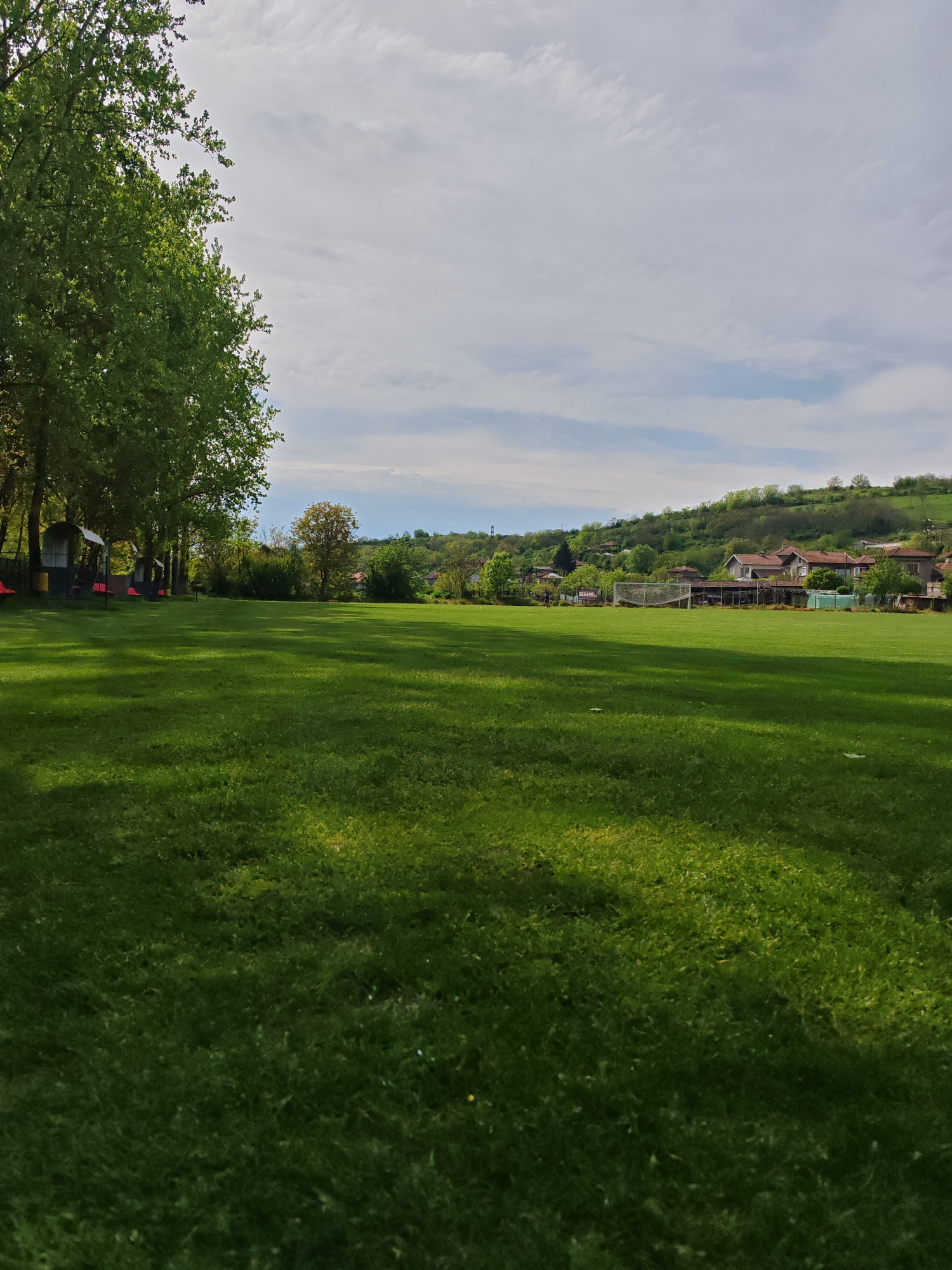 The image depicts a lush green field, likely a sports field or a large lawn, under a partly cloudy sky. On the left, a line of mature trees provides shade. Several sheltered benches are positioned beneath the trees. In the distance, a few houses are visible, situated on a gentle hillside. A soccer goal is observable in the middle distance. The location is possibly near Tsarevets, Bulgaria, based on the appearance of the architecture and landscape, but without additional context, the precise location is not conclusively determined. The image does not contain people or active interactions, and the general environment is open and grassy, with trees and residential buildings in the distance.