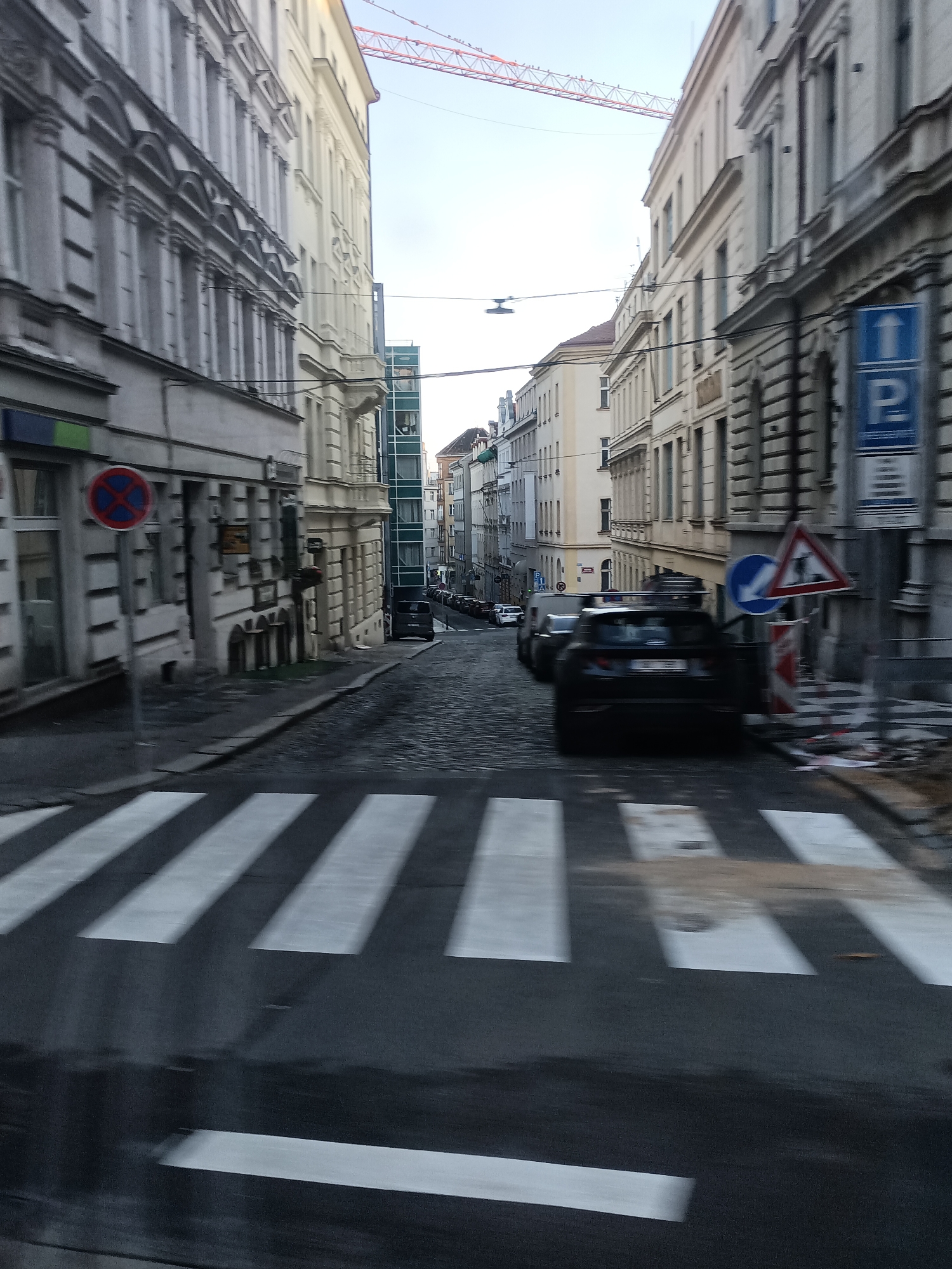 The image shows a street in Prague, Czechia, characterized by its traditional European architecture. On either side of the narrow, cobblestone street are multi-story buildings with ornate facades, typical of the city's historic districts. The buildings are a mix of light yellow and grey stone, with numerous windows and architectural details. A prominent feature of the scene is a construction crane visible in the background, suggesting ongoing development or renovation in the area. Traffic signs are present, including a "no parking" sign and road signs indicating direction and parking. Several cars are visible on the street, including a dark SUV in the foreground and other vehicles further down the road. The street has a pedestrian crossing marked with white stripes. The overall impression is a view of a historic European city street with a blend of old-world charm and modern activity.