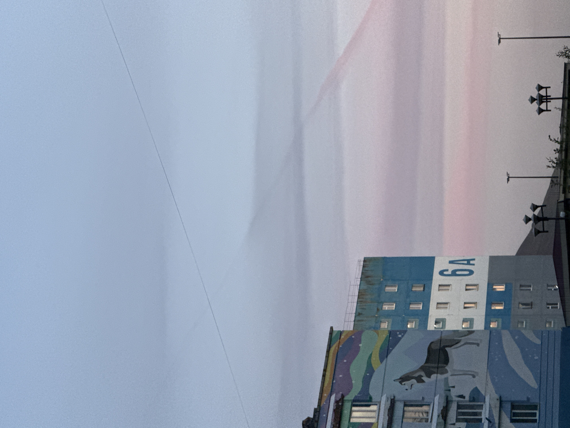 This image depicts a residential building in Norilsk, Russia, characterized by a mural on its facade. The mural features a husky dog and abstract, colorful wavy patterns, with snow and stars. The building itself is blue and white, with numerous windows. The number "6A" is prominently displayed on the building's side. The sky is a soft gradient of blue and pink, suggesting either dawn or dusk, with a thin wire stretching across it and a faint contrail from an aircraft visible. The lower portion of the image shows streetlights and the top of another building. There are no people visible in the scene.