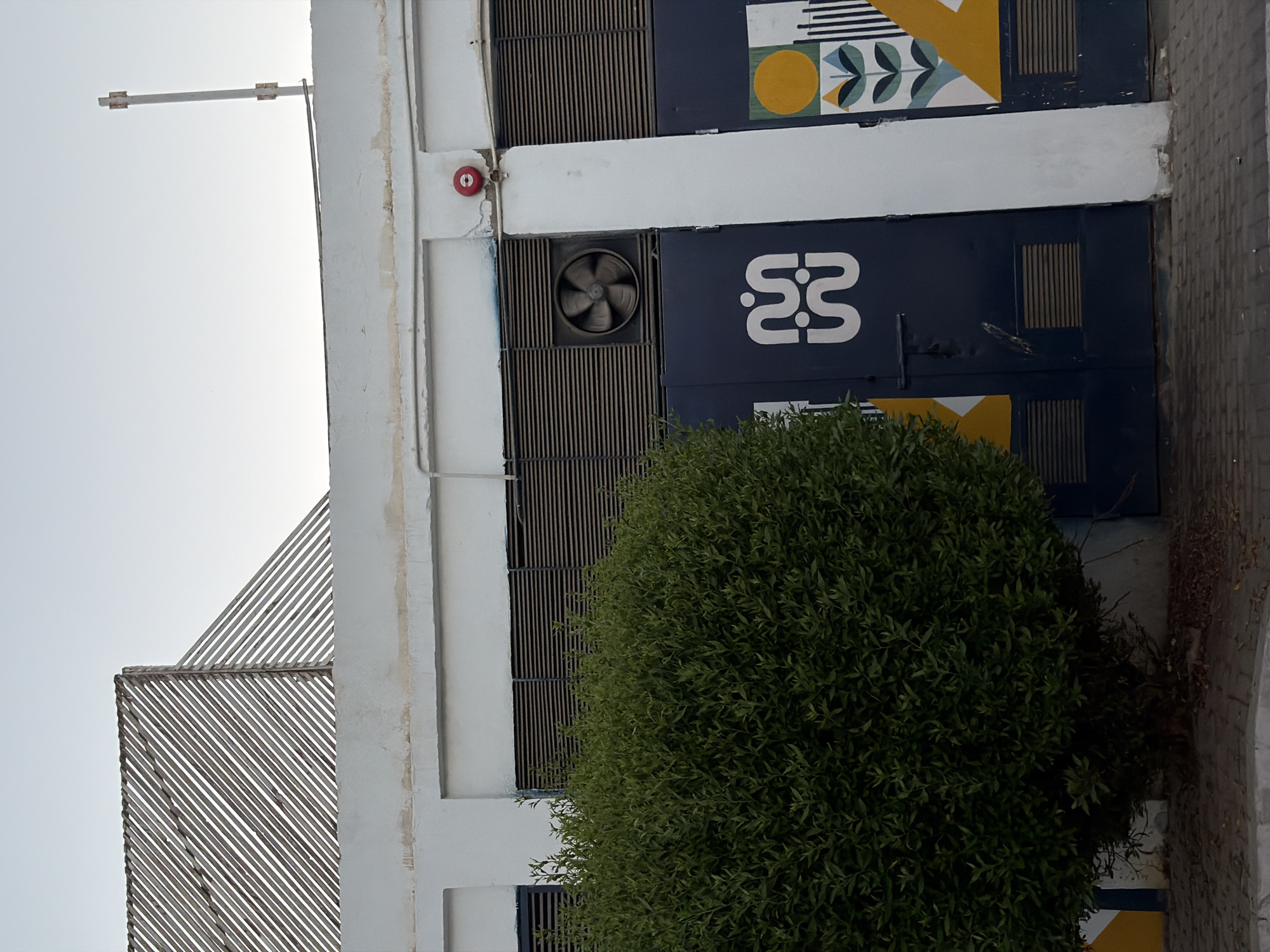 The image shows a close-up view of a building facade with some architectural and decorative elements. The building is white with dark blue panels and a geometric patterned mural featuring circles and stylized leaves. A circular red fire alarm is visible on the white section of the wall. Below this, a square vent with a fan is set into a slatted dark panel. A logo, appearing as two stylized "S" shapes, is printed on one of the dark blue panels.
To the left of the building, a diagonal, slatted structure made of thin beams is visible, suggesting an awning or shade element. A tall, dense green bush occupies the foreground, obscuring the lower part of the building and extending to the right. The sky is a pale, overcast gray, indicating a daytime scene, possibly with cloudy weather. The overall impression is of an exterior urban or commercial setting, perhaps in a location like Sharm El Sheikh, Egypt, given the architectural style and climate. No people are visible in the image.