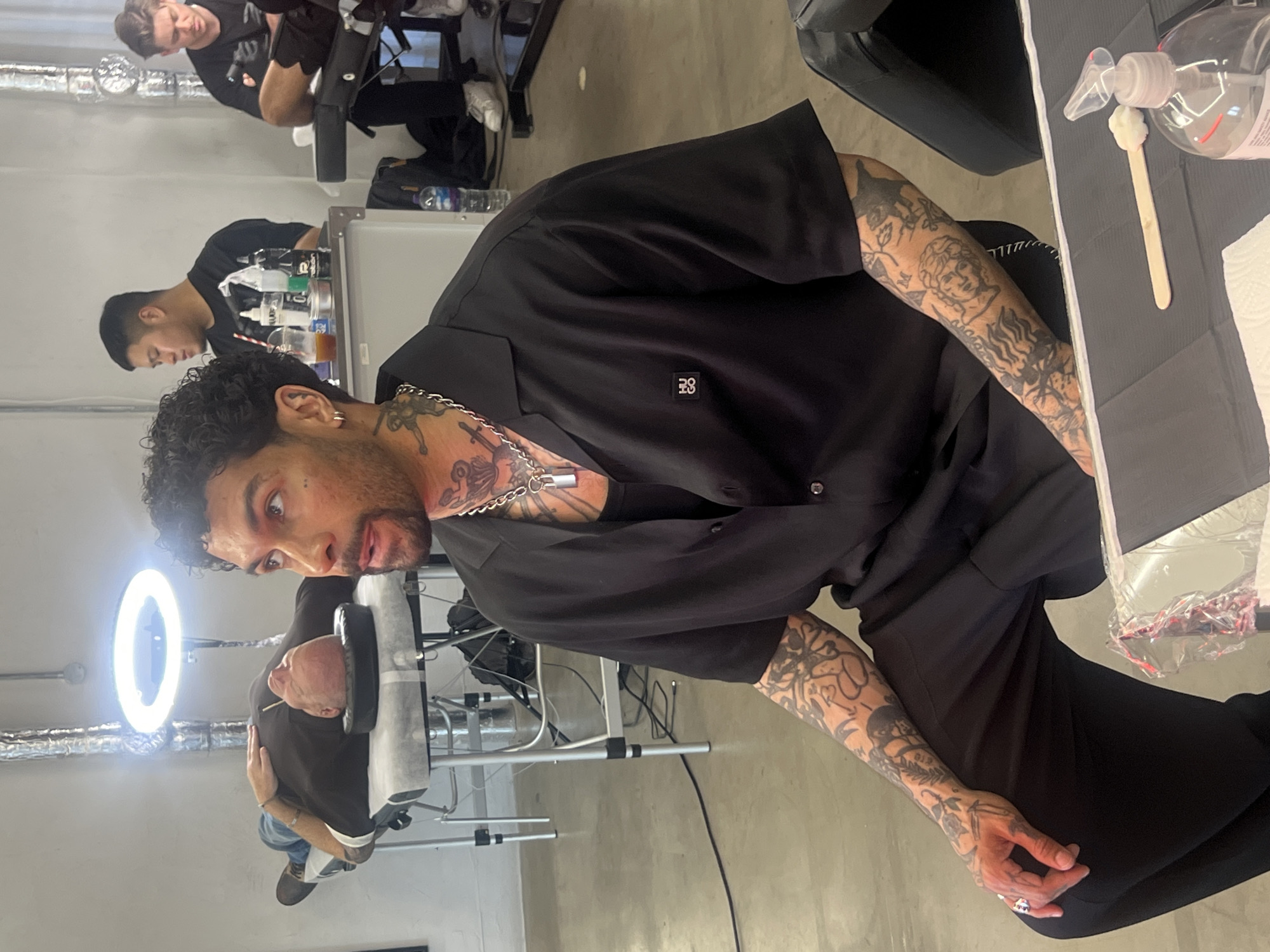The image captures an indoor scene, likely a professional tattoo studio. The main subject is a man with curly dark hair and a beard, looking directly at the camera with an intense expression. He is heavily tattooed on his arms, neck, and chest, featuring intricate designs including classical faces and botanical elements. He wears a black short-sleeved shirt with a small, square, dark emblem on the left chest, a silver chain, and rings.
In the background, another man lies face-down on a black tattooing table, his head resting on a black headrest. A prominent ring light is visible near his head, providing illumination. Further back, two other men are present; one appears to be seated at a station with various bottles and supplies, suggesting he is also a tattoo artist or apprentice. The setting features a minimalist, industrial design with white walls, a concrete floor, and exposed metallic pipes on the ceiling.
On a work surface to the right, a clear bottle of hand sanitizer with a pump top, a white wooden stick, and paper towels are neatly arranged on a grey mat, emphasizing the hygienic nature of the environment. The overall activity depicted is consistent with a tattoo session or a break within a tattoo studio setting. The emblem on the main subject's shirt is too small to read clearly but appears to be a stylized logo or initials.