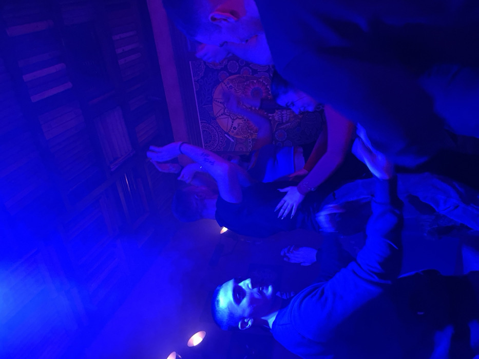 The image, rotated counter-clockwise, captures an indoor scene depicting several individuals under intense blue artificial lighting, suggesting a night event in a lively venue such as a club or bar in Drapetsona, Greece.
In the immediate foreground, a man in a dark top is visible, looking towards the left with a focused or intense expression. Centrally, another man, also in dark attire, is captured with his arms and hands raised in front of him, possibly mid-dance or performing a gesture. Behind him, a woman's hands are gently placed on his lower back, indicating interaction or partnered movement. Further in the background, two more individuals are partially visible, one looking down towards the central figures and another observing from behind the woman.
The setting includes a decorative background featuring a patterned wall hanging or tapestry with circular and floral designs in muted warm tones, visible despite the pervasive blue light. Adjacent to this, a wall section appears to be constructed from wooden panels or shelves. Several small, circular light sources are positioned on what seems to be the floor or a low surface, emitting warmer light that contrasts with the scene's dominant cool blue hue. A slight atmospheric haze or smoke is present, contributing to the ambiance.
The activity appears to be a social gathering, possibly a dance performance, party, or a lively night out, characterized by movement and interaction among the participants. The expressions of the individuals are largely obscured by the lighting or their orientation. No visible text is present in the media file.