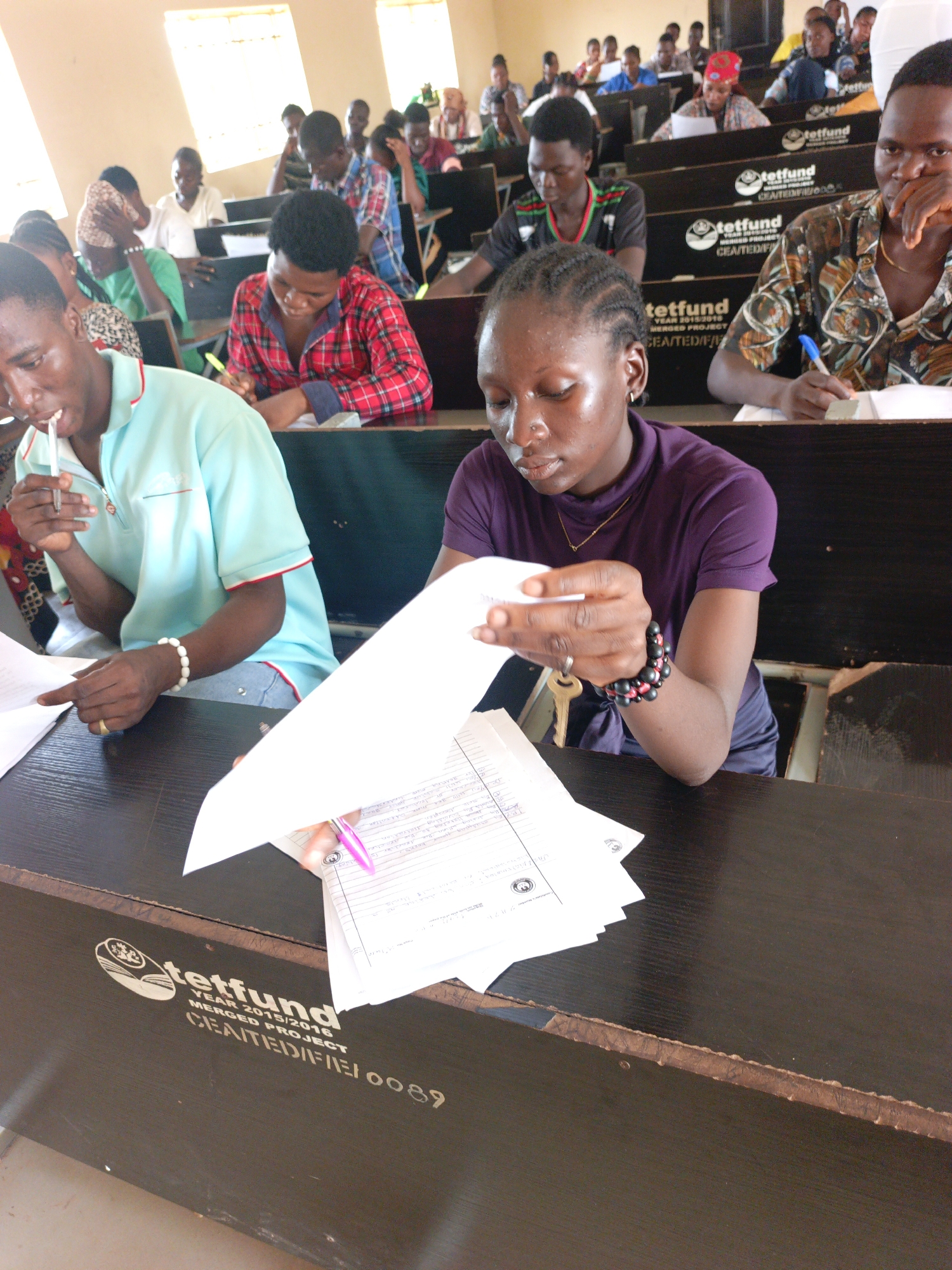 The photograph shows a classroom full of students seated at rows of desks. The students are predominantly young adults, with various skin tones and hairstyles. They appear to be engaged in some sort of academic activity, as most have papers and pens and are focused on their desks.
The desks are made of dark wood and have logos printed on them, indicating that they were funded by "tetfund" for a project spanning the years 2015/2016. One logo explicitly mentions "CEA/TEDIFIERI 0089" and other mentions "CEA/TEINFIRE000." This clue suggests the image may have been taken in Akwanga, Nigeria, as tetfund is a Nigerian government agency dedicated to funding educational projects.
The environment looks like a typical lecture hall, with rows of desks facing forward, indicating a traditional classroom setup. The lighting appears natural, coming from windows that are visible in the background.
Overall, the scene depicts a typical academic environment where students are engaged in their studies.