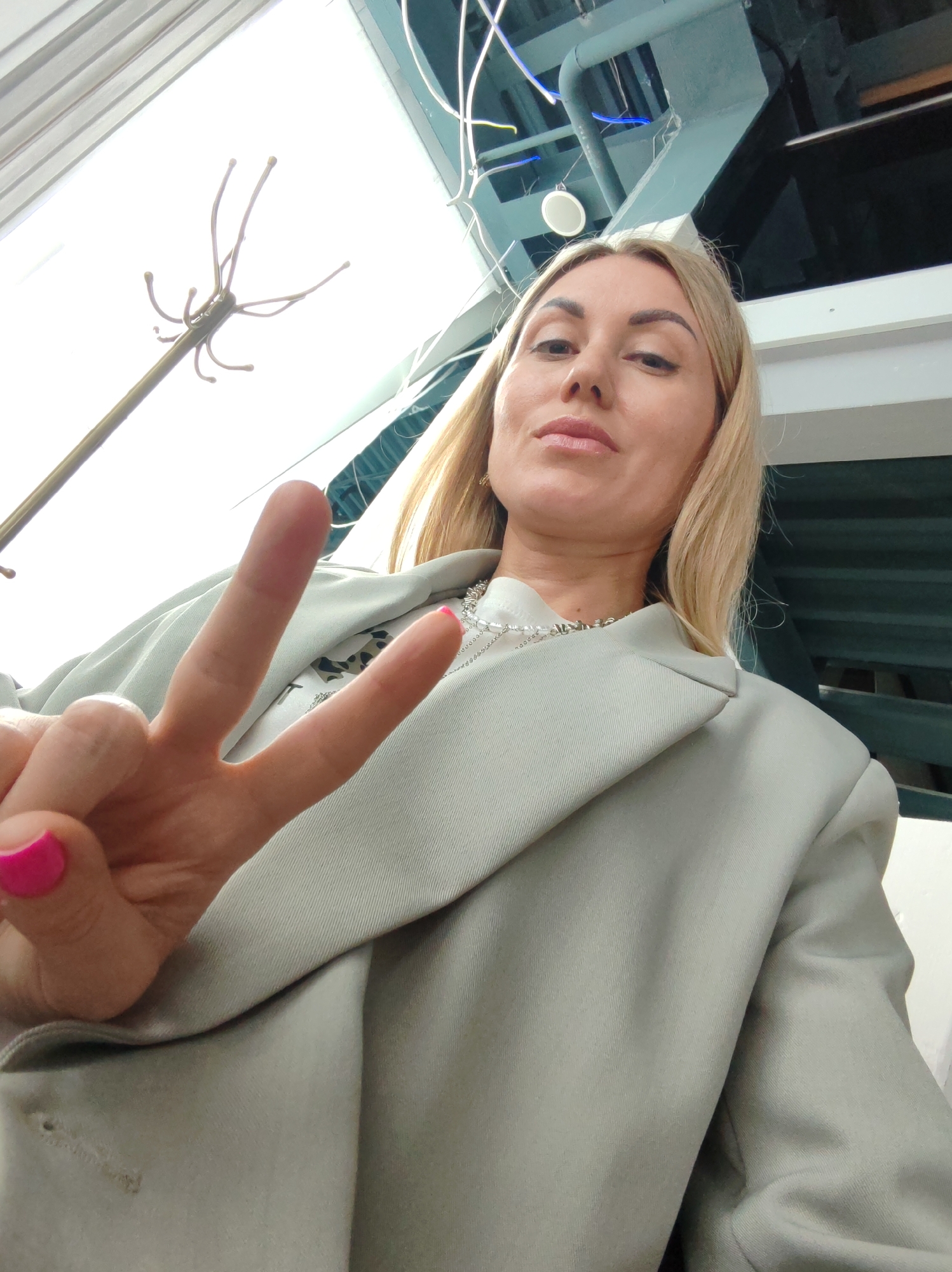 In a close-up, low-angle indoor shot, a blonde woman with light makeup, wearing a light gray blazer and a silver necklace, holds up two fingers in a peace sign. Her fingernails are painted bright pink. The background features a stark white ceiling with visible industrial elements like pipes and wires, and dark green horizontal blinds. A coat rack with multiple hooks is visible to the left. The setting does not provide any identifiable landmarks or characteristics specific to the city of Amursk, Russia.