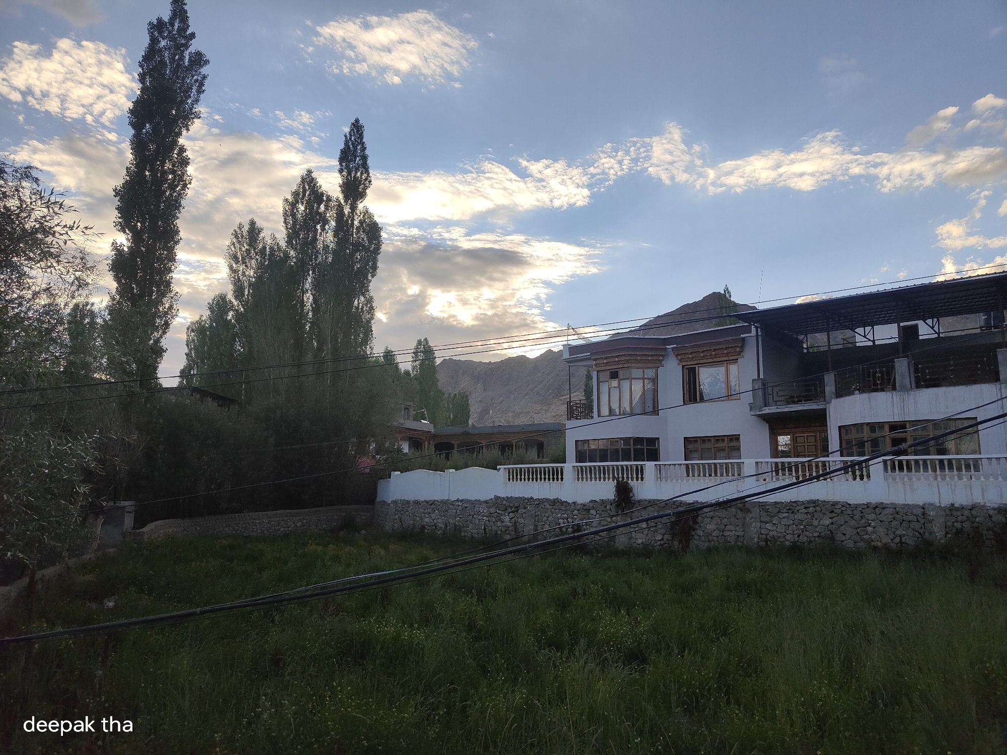 The image depicts a modern multi-story building with a white facade and wooden window frames, situated in Leh, India. The building is characterized by its balconies, a prominent rooftop structure, and is partially obscured by trees. A low stone wall with a white decorative railing separates the building from a grassy area in the foreground. Electrical wires run across the scene, both in front of and behind the building. The background features a mountain range under a sky with scattered clouds. The overall atmosphere suggests a rural or semi-urban setting in Leh. The lower-left corner of the image contains the text "deepak tha".