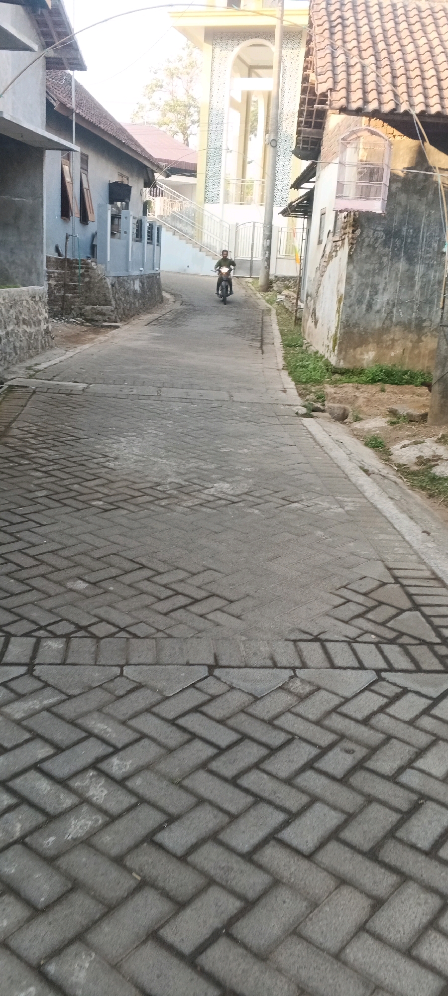 A person is riding a motorcycle down a narrow street in Penanggungan, Indonesia. The street is paved with interlocking gray bricks and is lined with modest houses on either side. In the background, a mosque with a white minaret and yellow accents is visible, along with some trees and a bright sky. The overall scene depicts a quiet, residential street in a rural or semi-urban area.