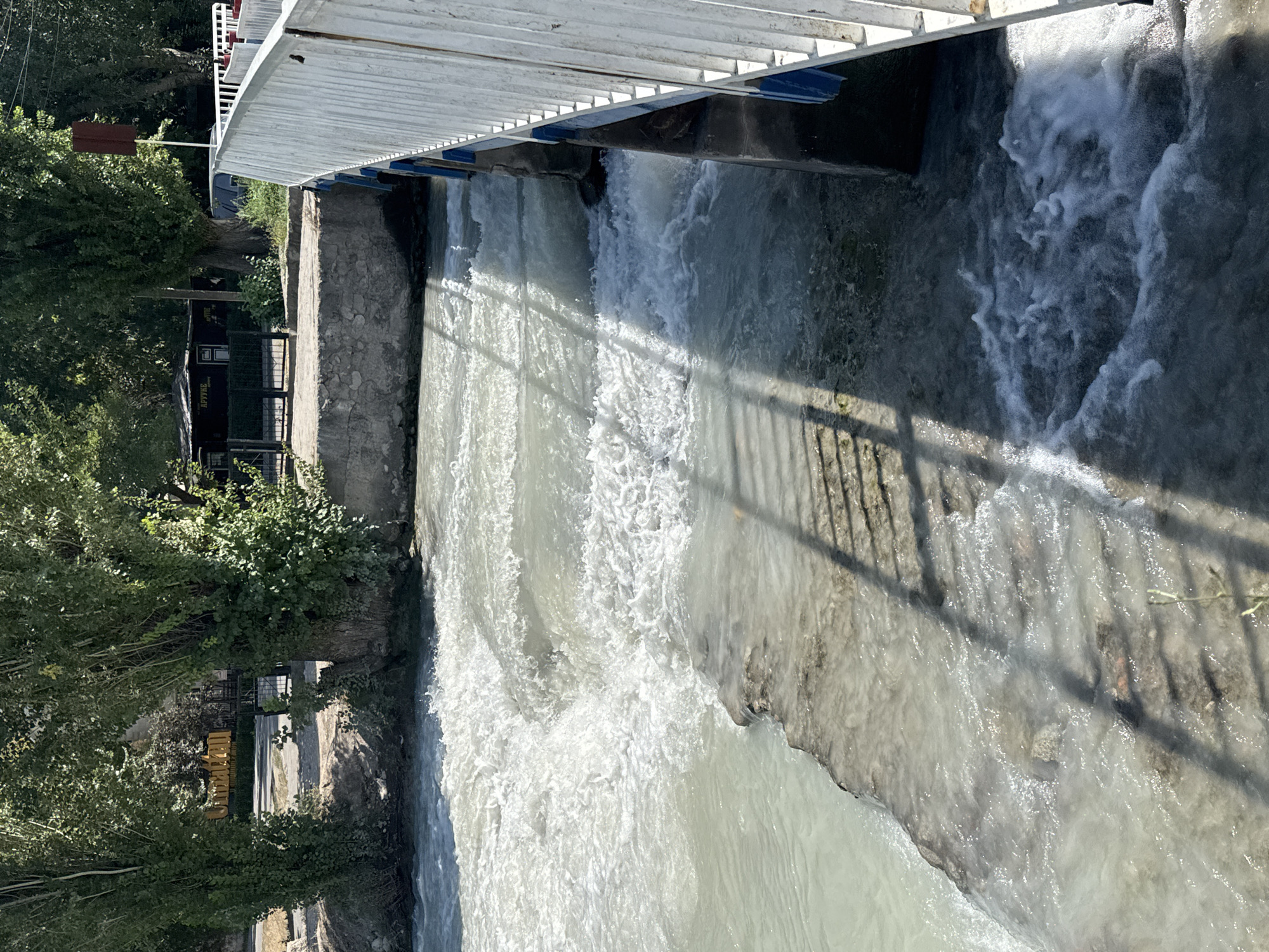 The image depicts a section of the Ala-Too River in Bishkek, Kyrgyzstan, flowing under a pedestrian bridge. The water is turbulent, with visible rapids and foam, suggesting a fast current. The bridge, constructed with white metal railings and blue accents, casts a shadow onto the water. The riverbanks are lined with concrete and stone structures, and lush green trees frame the scene. In the background, a small building with signage and a fence can be seen, hinting at a park or recreational area. The overall impression is one of a natural environment integrated into an urban setting.
