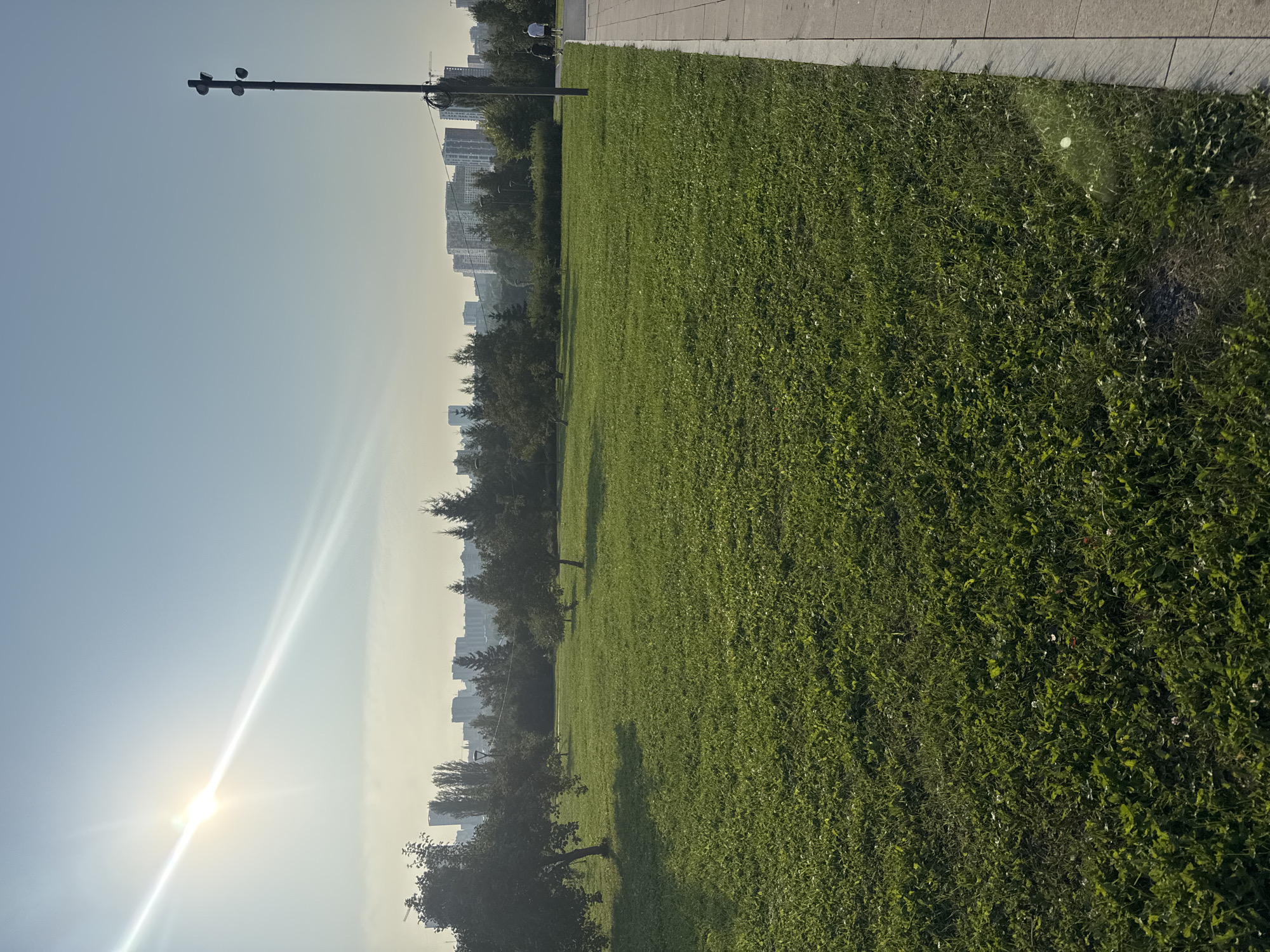 The image depicts a park area in Ufa, Russia, on a clear day. A wide expanse of well-maintained green grass occupies the foreground and mid-ground. Scattered trees are visible on the left side, casting shadows on the grass. In the distance, a city skyline with various buildings and skyscrapers is present. The sky is bright blue with the sun prominently featured in the upper left quadrant, creating a lens flare that extends diagonally across the sky. A paved walkway runs along the right edge of the frame, with two individuals walking on it. A tall lamppost with multiple light fixtures stands near the walkway. The overall scene suggests a recreational area within an urban setting.