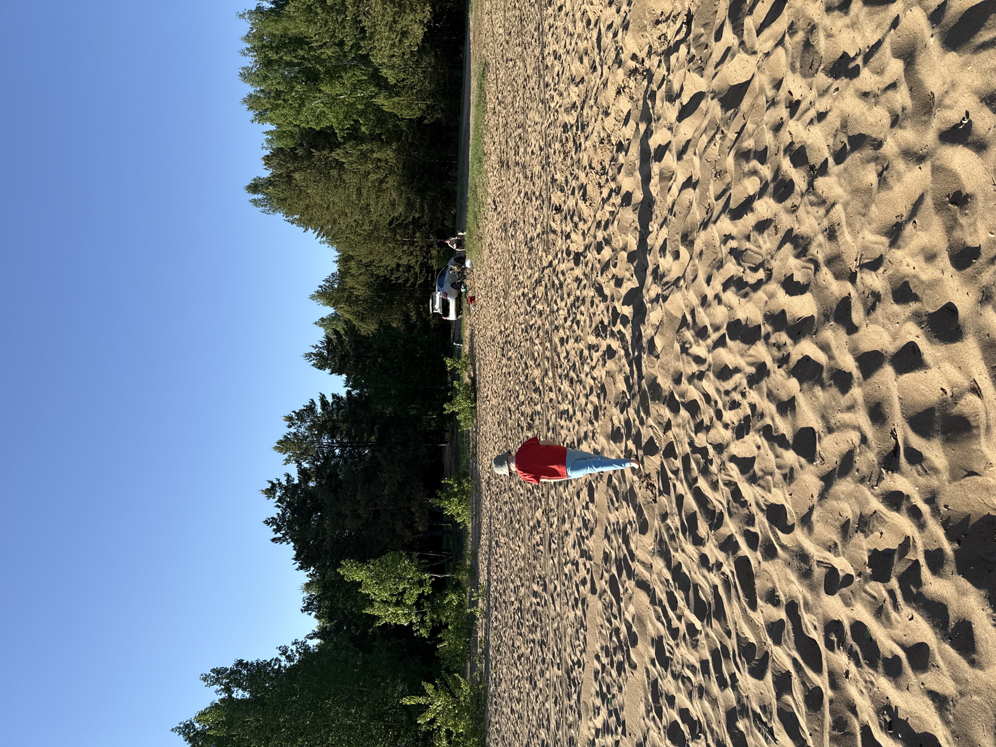 A daytime, wide-angle, outdoor shot shows a sandy area adjacent to a tree line with a person lying on their stomach in the sand. The person is wearing a red shirt, blue pants, and a light-colored hat. The trees are green and dense. The sky is clear and blue. In the distance, a white vehicle is visible, and two people stand near the vehicle. The sand is textured with shadows, possibly caused by footprints and wind patterns.
The location is Noviy, Russia.
