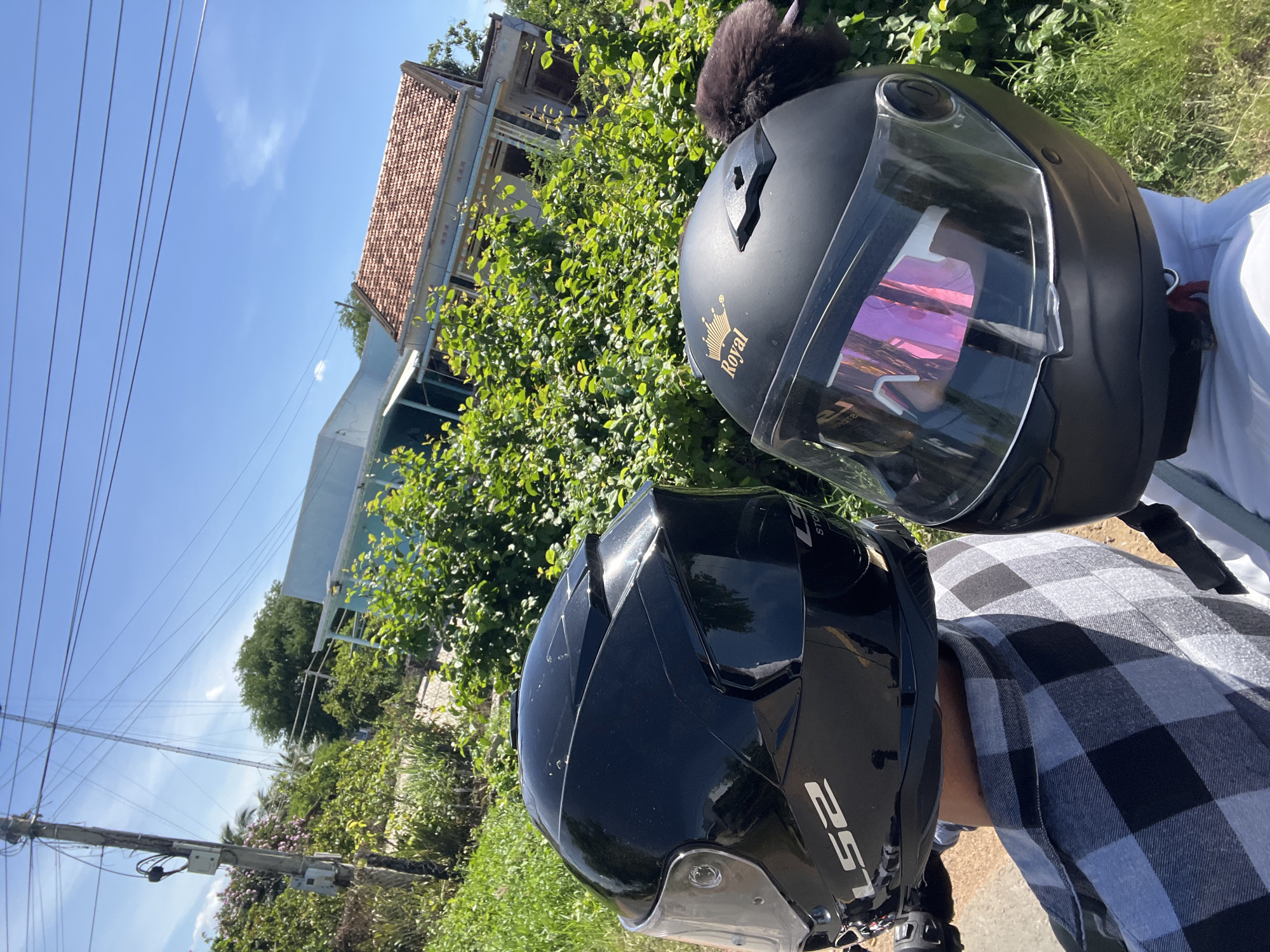 Two individuals wearing helmets are seated on a vehicle, facing forward, with a rural landscape in the background. The person on the left wears a black LS2 helmet and a grey and black checkered shirt. The person on the right wears a black helmet with the brand name "Royal" in gold script and a pink-tinted visor. They are positioned in front of dense green foliage and a light blue building with a red-tiled roof. Power lines are visible against a clear blue sky, and a utility pole stands to the left. The location is identified as Hong Son, Vietnam.
