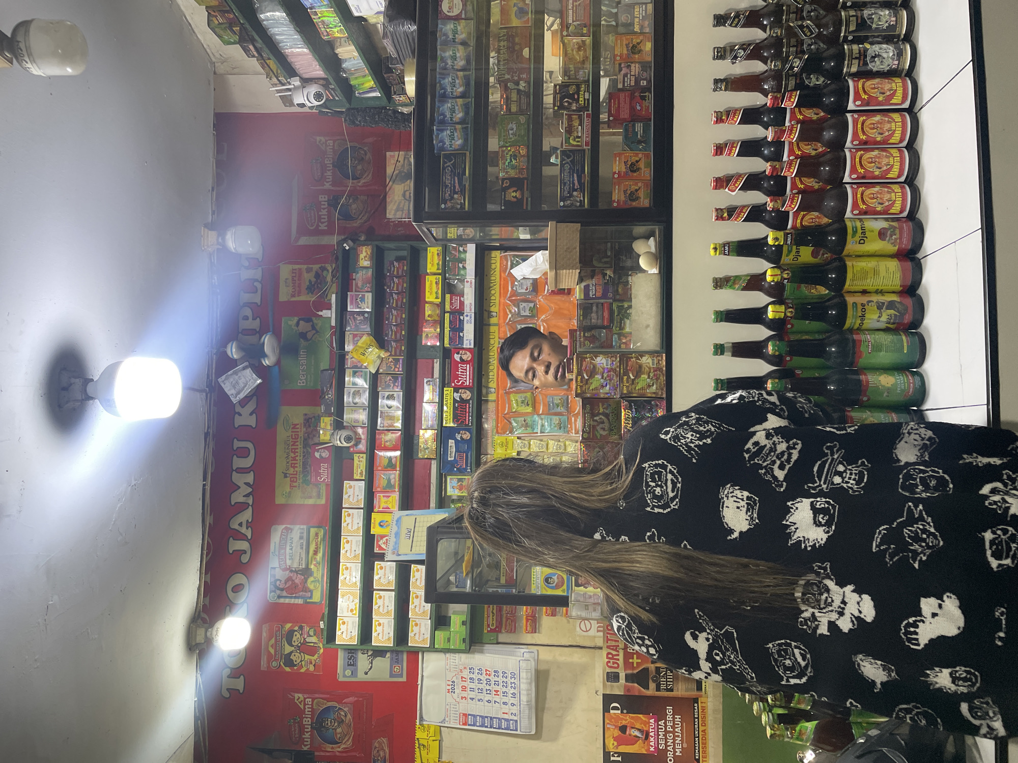 The image depicts the interior of a "Toko Jamu" (traditional herbal medicine shop) located in North Jakarta, Indonesia. A customer, with long light brown hair, is standing in front of a counter, partially obscuring the view. Behind the counter, a shopkeeper in an orange shirt is visible. The shop is densely stocked with various items, including rows of bottled herbal medicines, packaged goods displayed on shelves, and promotional posters on the walls. A calendar dated May 2026 is hanging on the wall near the counter. Lighting fixtures are visible on the ceiling. The camera angle is slightly tilted, adding to the close-quarters feel of the shop.