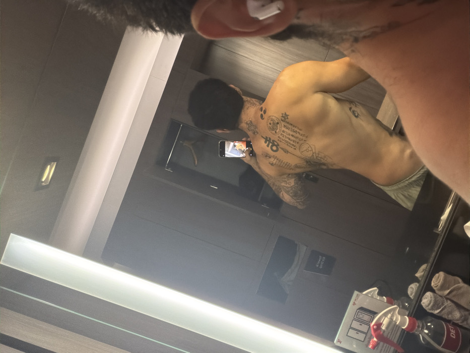 A male individual is observed taking a mirror selfie in a hotel room in Bangkok, Thailand. The subject is reflected from the back, shirtless, revealing extensive tattoos across his entire back, shoulders, and arms, featuring traditional Sak Yant style and symbolic designs. He is wearing light-colored shorts or underwear. He holds a dark-cased smartphone, whose screen reflects a blurred, colorful image. In the immediate foreground, the right side of the photographer's head, including an ear with a white earbud, and a tattooed portion of the neck and shoulder are visible. The reflected room environment displays modern dark wood paneling, a wardrobe section with a single wooden hanger, and multiple linear and recessed lighting fixtures. On a dark vanity surface below the mirror, two spray bottles (one labeled "DETOX"), two rolled towels, a small white box, and a clear plastic sign bearing the text "KRUNG THEP" are arranged.