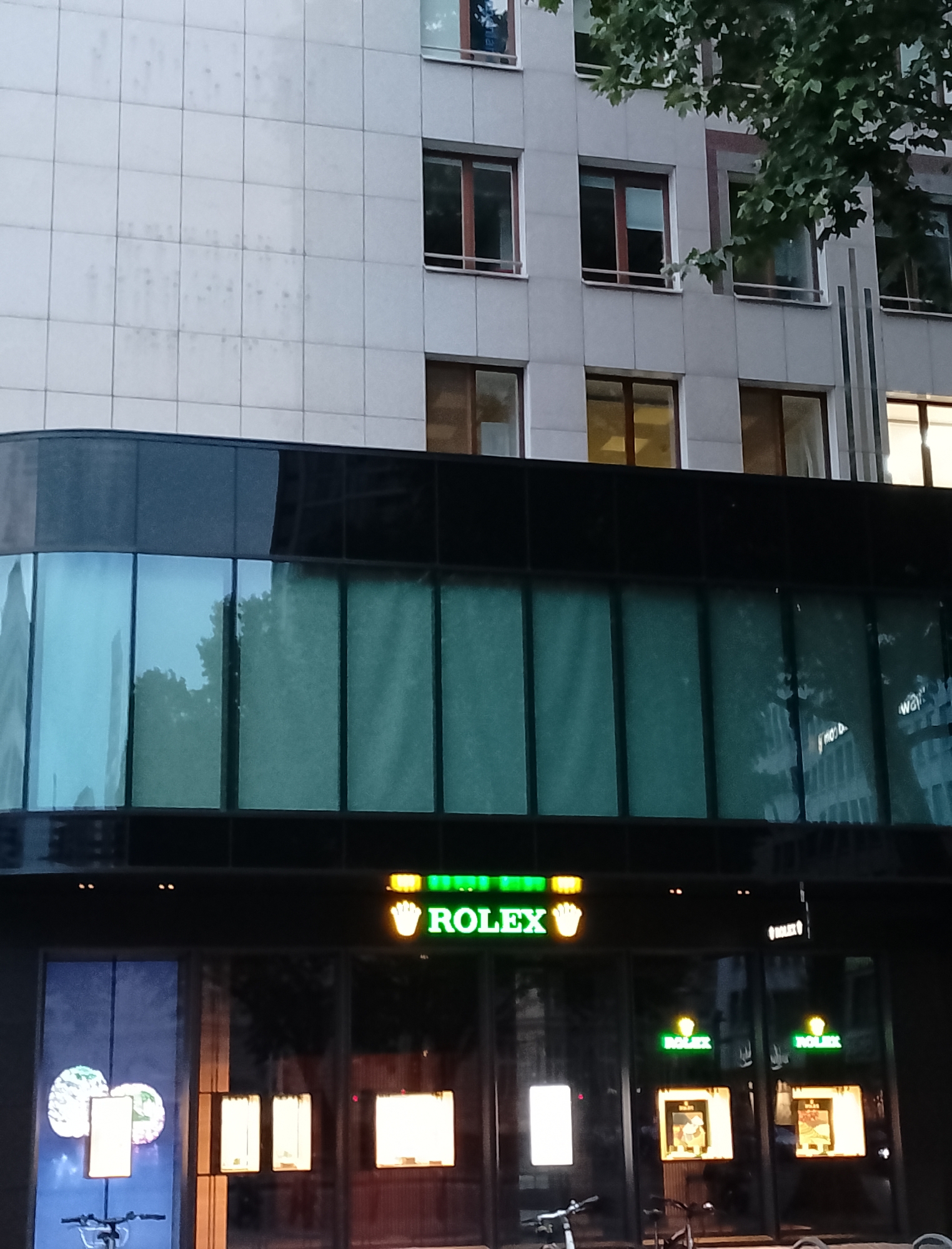 A medium shot captures the facade of a Rolex store in an urban setting, likely in the city of Unknown. The building features a modern architectural style with a light-colored tiled exterior on the upper floors and a sleek, dark facade with large glass windows on the lower level. Green illuminated signs with the "ROLEX" logo are prominently displayed above the entrance, accompanied by the iconic Rolex crown symbol in gold. Inside the store, illuminated display cases showcasing watches are visible through the glass. The reflections in the windows suggest the presence of trees and other buildings across the street. A bicycle is parked in front of the store on the left side of the frame. The overall impression is one of a high-end retail establishment in a contemporary urban environment.