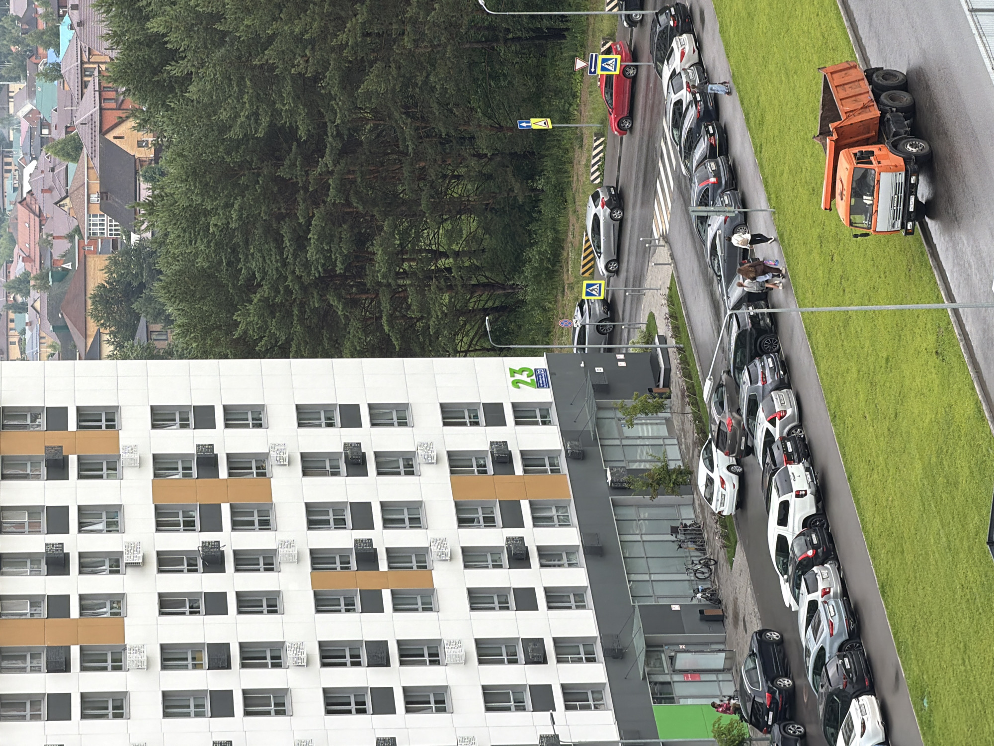 The image shows an aerial view of a street scene with a modern apartment building on the left and a row of parked cars on the right, bordered by a grassy area. The apartment building is white with a repeating pattern of windows and some orange-colored accents. The cars are of various makes and models, parked in a line along the street.
On the right side of the image, an orange dump truck is parked, its bed tilted up. A few people are visible near the dump truck, and some are walking along the sidewalk. There are also several bicycles parked near the apartment building.
Road signs are visible in the background, including a yellow pedestrian crossing sign. The sky appears overcast, suggesting a cloudy day. The overall setting is a residential or mixed-use area with modern architecture and green spaces. The image is oriented vertically, with the top of the apartment building appearing at the bottom of the frame.
Visible text includes "23" in green on the apartment building.