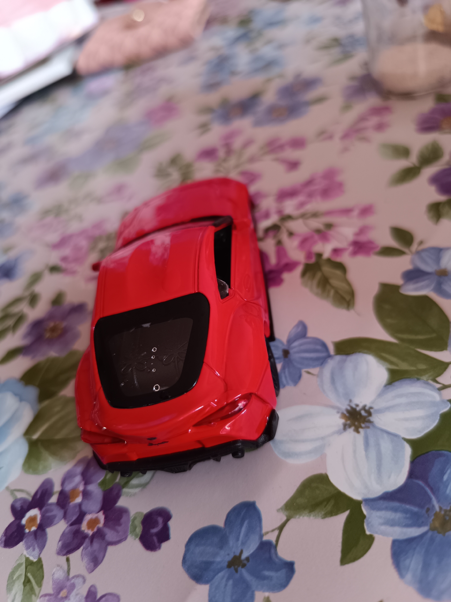A red toy car sits on a tablecloth with a floral pattern. The car has a black rear window with some white circles visible inside. The background is out of focus, but there is a glass jar and some other objects on the table. The location is unknown.