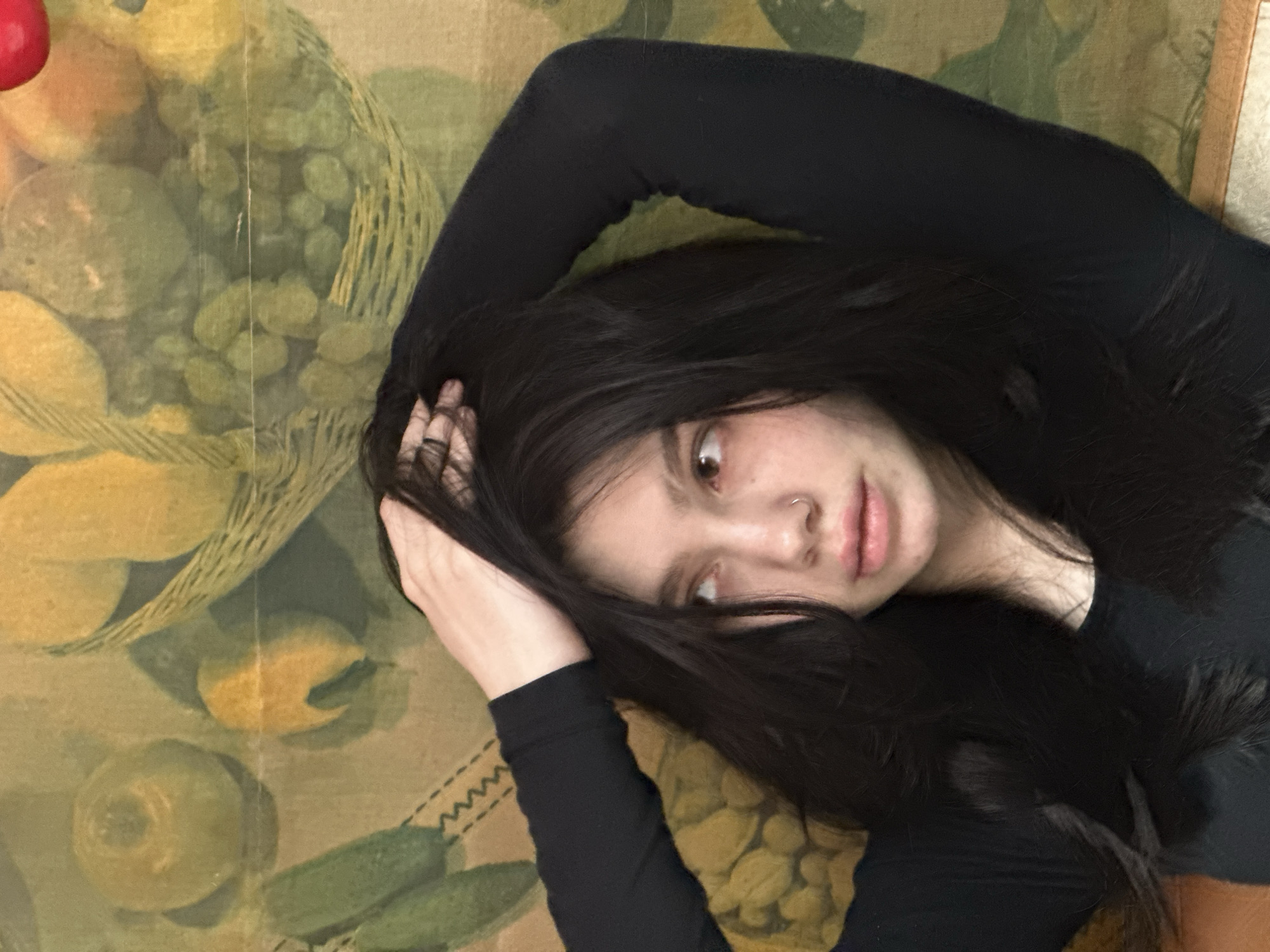 A close-up vertical photograph taken in Aktanysh, Russia, depicts a young woman from the chest up, positioned against a patterned backdrop. The woman has long, dark hair which she holds with both hands intertwined above her head. She wears a plain black long-sleeved garment. Her face is turned slightly to the left, with her gaze directed upwards and to the right, away from the camera. A silver-colored ring pierces her left nostril. The background is a fabric or surface displaying muted green, yellow, and brown illustrations of fruits and vegetables, including a round green fruit, a yellow pear, elongated green vegetables, and a basket containing small oval fruits. A prominent horizontal crease is visible across the mid-section of the background.