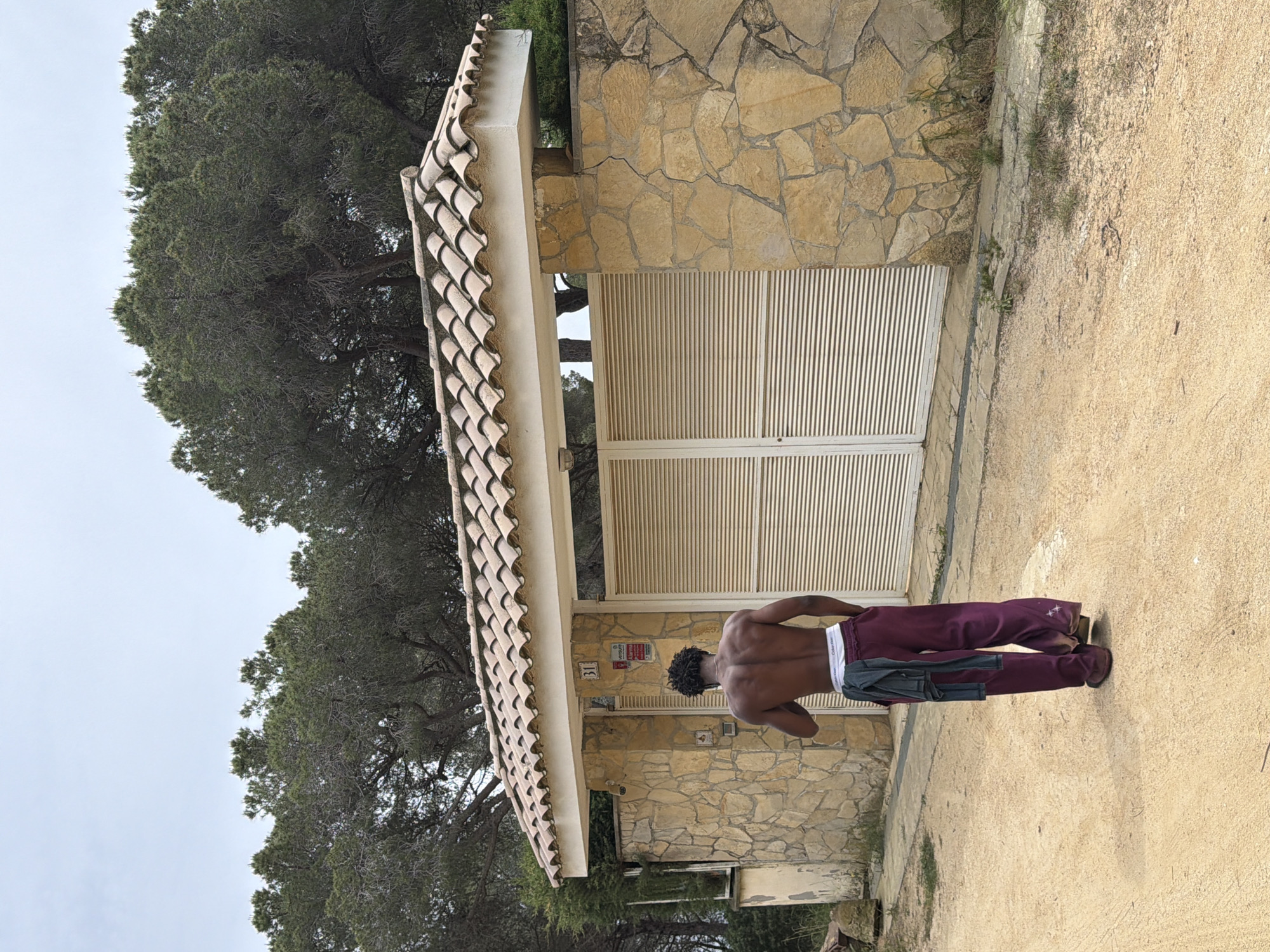 A shirtless man wearing maroon sweatpants stands facing a building in Sant Andreu de Llavaneres, Spain. The building is constructed of irregular light-colored stone with a tiled roof and white louvered doors. Trees are visible behind the building. The ground is a mix of sand and sparse vegetation.