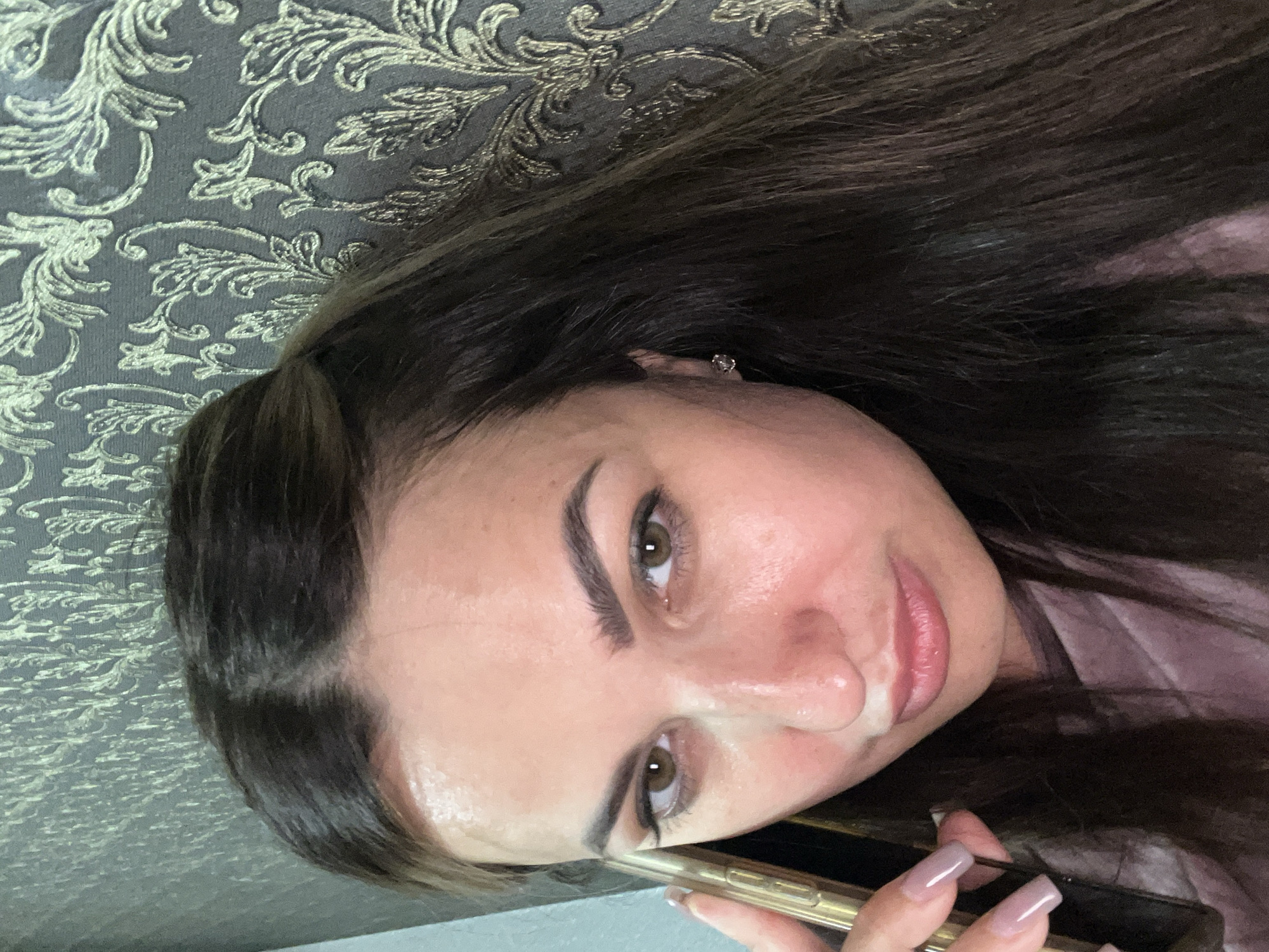 A woman, with dark brown hair and light brown eyes, is shown in a close-up, indoor shot. She is holding a gold-colored smartphone to her right ear, as if on a call. Her left hand, with manicured nails, is near the phone. She is wearing a pinkish-purple top. The background features a textured wallpaper with an ornate, repeating floral pattern in a metallic gold and grey color scheme. The lighting appears to be soft, with a slight sheen on her forehead and nose, suggesting some sweat or makeup. Her expression is neutral, with a slight tilt of her head and a furrowed brow.