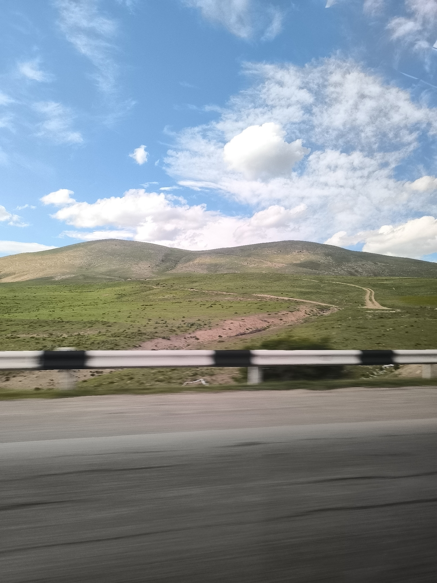 This image captures a vibrant, expansive landscape as seen from a moving vehicle in Yelpin, Armenia. The scene is dominated by undulating, green hills under a bright blue sky dotted with scattered white clouds, some appearing wispy while others are more defined cumulus.
In the foreground, a heavily motion-blurred asphalt road indicates travel, and a black and white striped metal guardrail runs across the mid-ground, also exhibiting motion blur. Beyond the guardrail, the rolling hills rise, covered primarily in lush green grass, with some patches of sparser, drier-looking vegetation. Several unpaved dirt tracks or paths can be seen winding through the verdant terrain of the hills, suggesting routes for off-road vehicles or hikers.
The time of day appears to be late morning or early afternoon, characterized by bright, even daylight. The weather is clear and sunny with pleasant cloud cover. The overall impression is one of a vast, natural, and relatively unpopulated mountainous region. There are no people or discernible text visible in the image. The image conveys a sense of a journey through scenic, rugged natural beauty.