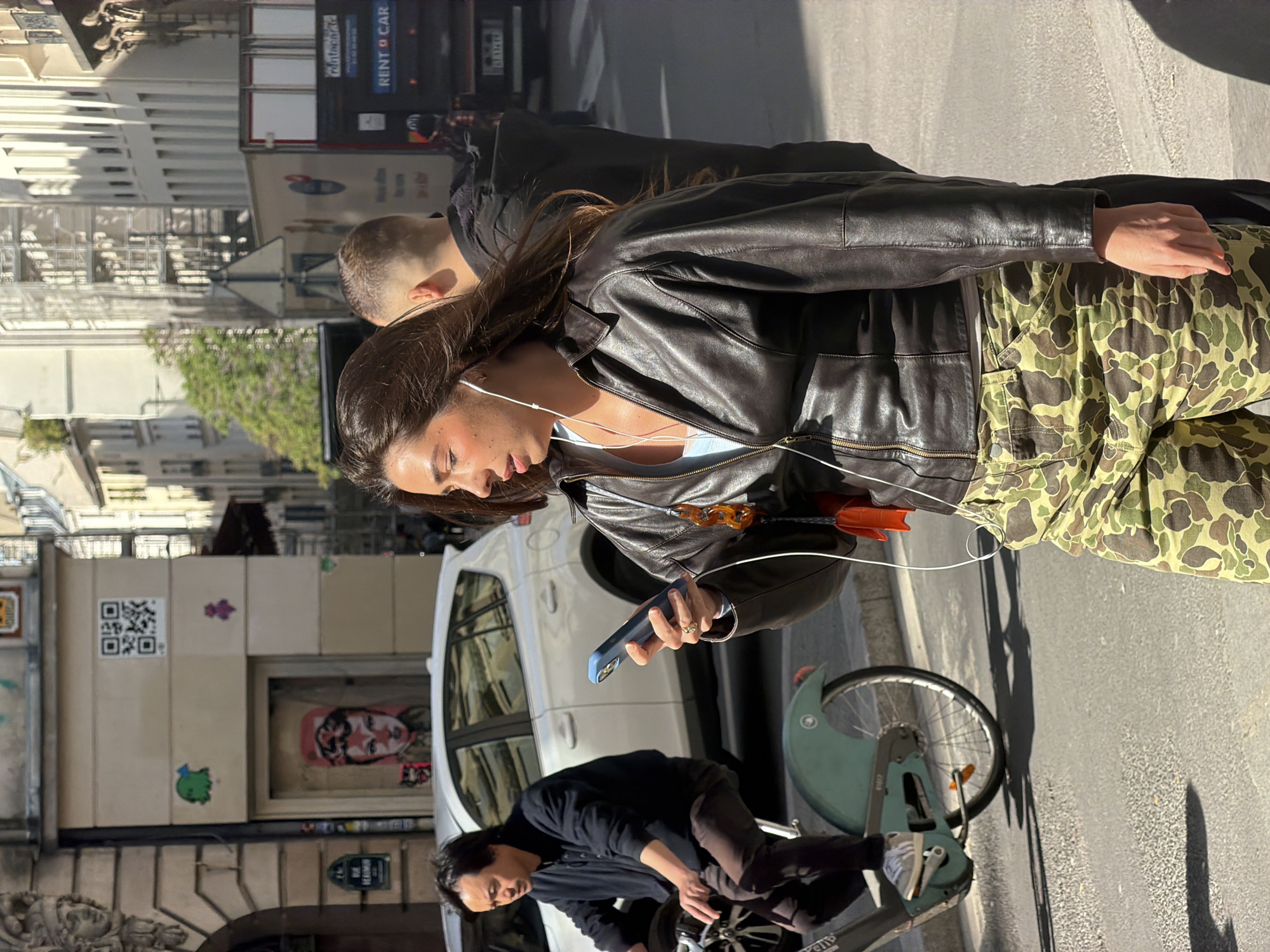 A woman wearing a black leather jacket and camouflage pants is standing on a street in Paris, France, holding a blue smartphone in her left hand. She has earphones in her ears. Next to her is a man's head with short hair, and a second man is crouched on a green bicycle. A white car is parked behind them. Buildings line the street, one with a QR code and a graffiti image of a woman's face on the wall. A "RENT A CAR" truck is parked further down the street.