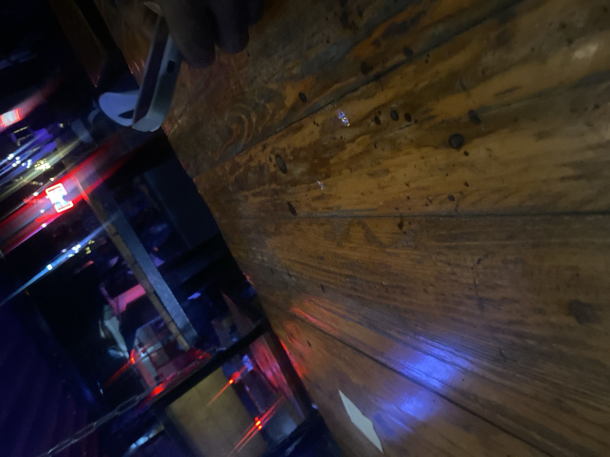 This image depicts an indoor establishment, likely a bar or nightclub, located in Howayi, Nigeria. The foreground features a wooden table, characterized by visible grain, knots, and dark, irregular marks, which exhibits reflections of prominent blue and red light sources. A person's hand, appearing with a darker skin tone, holds a white smartphone, partially obscuring it, on the right side of the table. A small, irregularly shaped white object is present on the left side of the table. The background is out of focus, revealing a dimly lit environment with numerous colored lights, predominantly red and blue, along with some distant white lights. Indistinct human figures, mostly appearing as silhouettes, are visible throughout the blurred background, suggesting other individuals in the establishment. Reflections of red light are visible on what appears to be a dark structural element or reflective surface separating the foreground from the background. A white, possibly plastic, chair back is faintly discernible in the mid-right background.