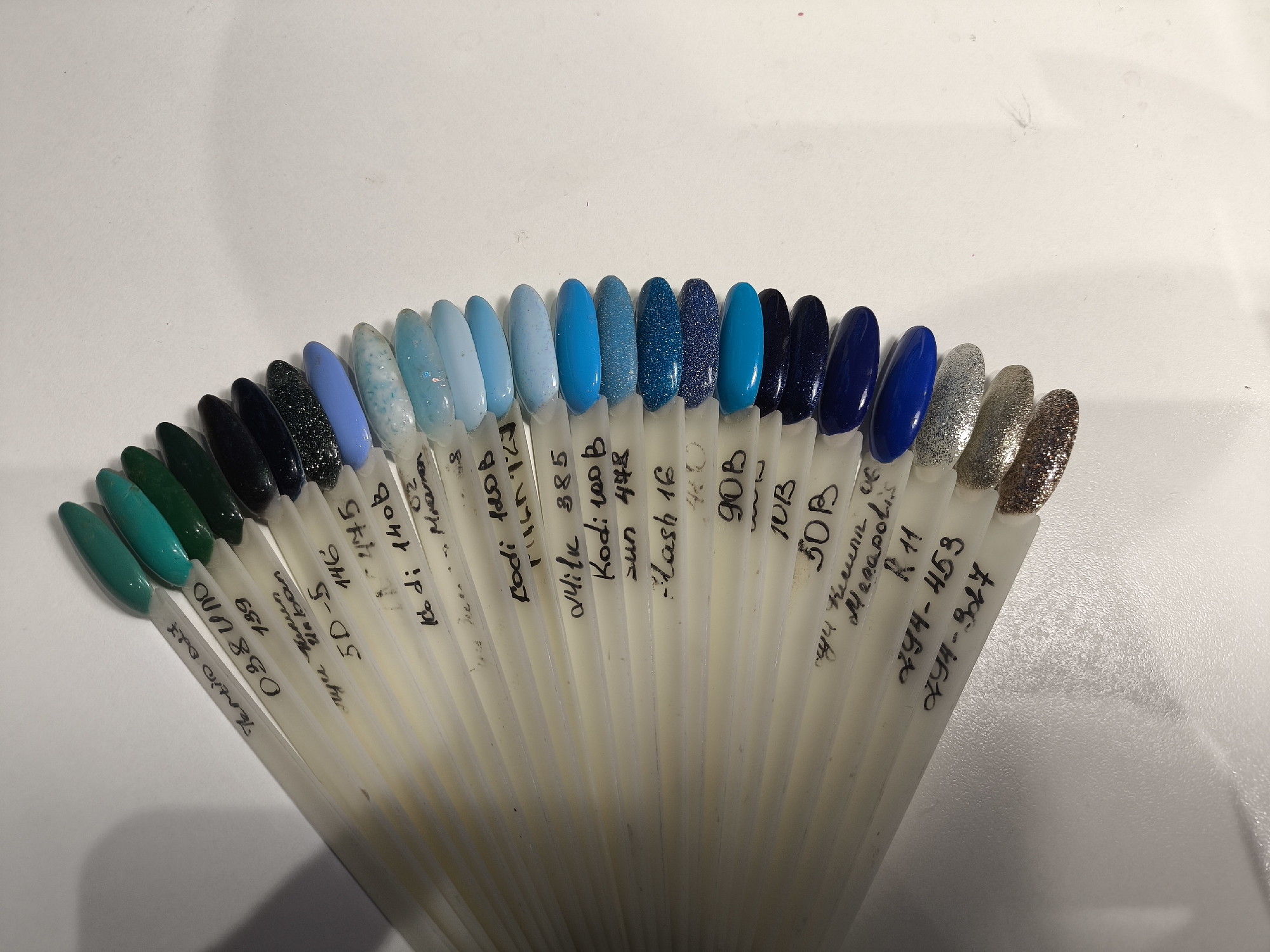 A collection of nail polish color swatches is fanned out on a plain white surface, likely a table or countertop, in an indoor setting in Khimki, Russia. The swatches are displayed on individual white plastic sticks, arranged in a spectrum primarily encompassing green and blue tones, with a few metallic/glitter shades at the end.
The colors transition from dark teal and emerald green on the far left, through various light blues, sky blues, medium blues, and deep navy blues. Some of the blue swatches feature speckled patterns or glitter finishes, while others are solid and glossy. Towards the right, the blues give way to shimmering metallic shades, including silver, gold, and bronze glitter. The lighting is even, suggesting an indoor environment.
Each plastic stick has handwritten labels in black marker, identifying the brand or color code of the polish. Visible text includes: "Tentio 047", "850", "657", "139", "Urban", "5D-5", "146", "445", "Kodi 140B", "Marmara 02", "Kodi 120B", "Milan", "Milky 385", "Kodi 100B", "Sun 448", "Flash 16", "90B", "10B", "50B", "My in Megapolis", "R11", "Z94-453", and "Z94-347".