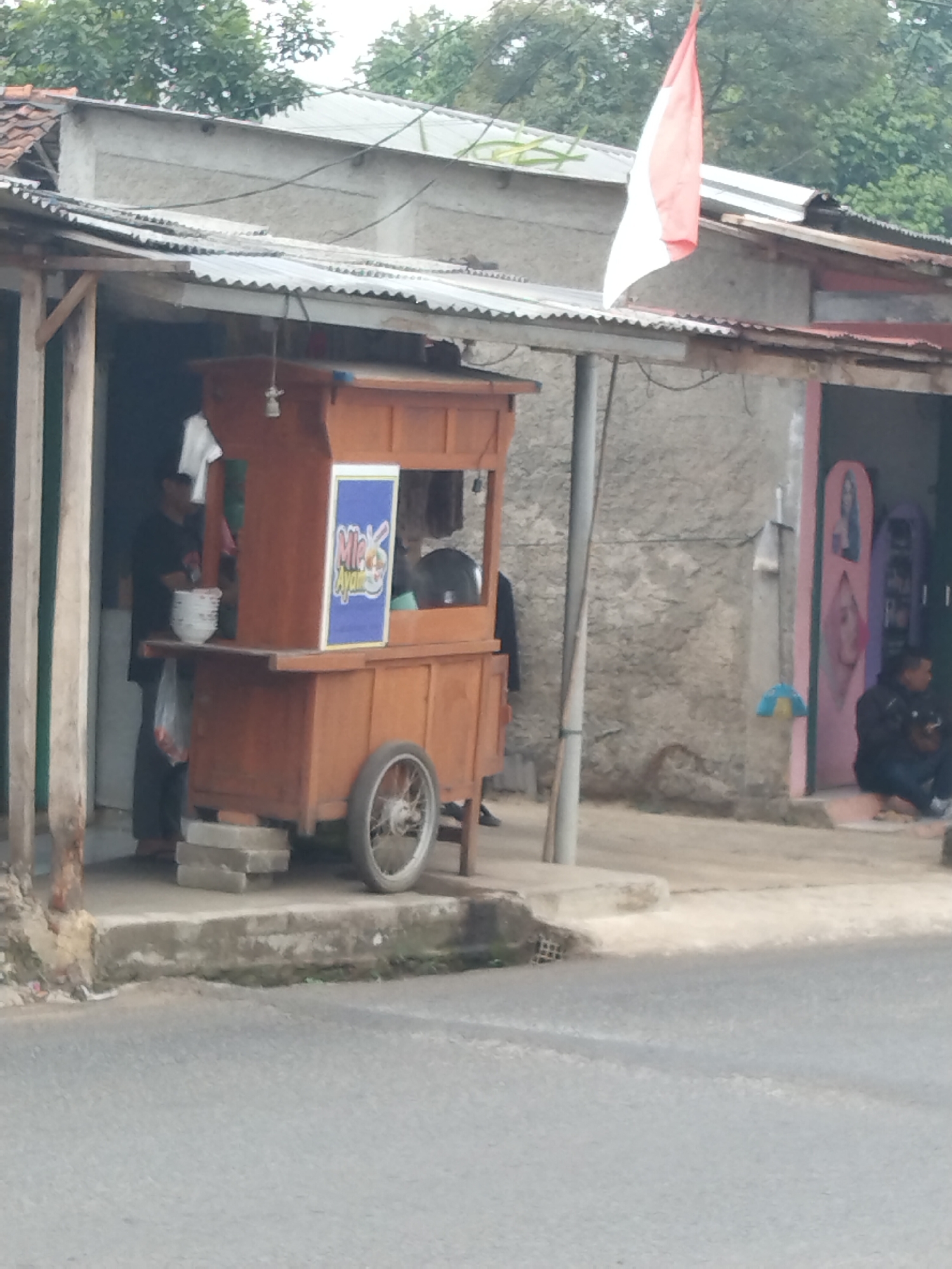 In Ciseeng, Indonesia, a food cart is parked on a street in front of a weathered building. The cart is made of wood and has a red and white sign that reads "Mie Ayam" in blue lettering. A person is standing behind the cart, and another person is seated in front of a shop to the right. The shop has colorful displays and a person is visible inside. A red and white Indonesian flag is flying from the roof of the building. The street appears to be made of asphalt, and there are some plants growing on the side of the road.