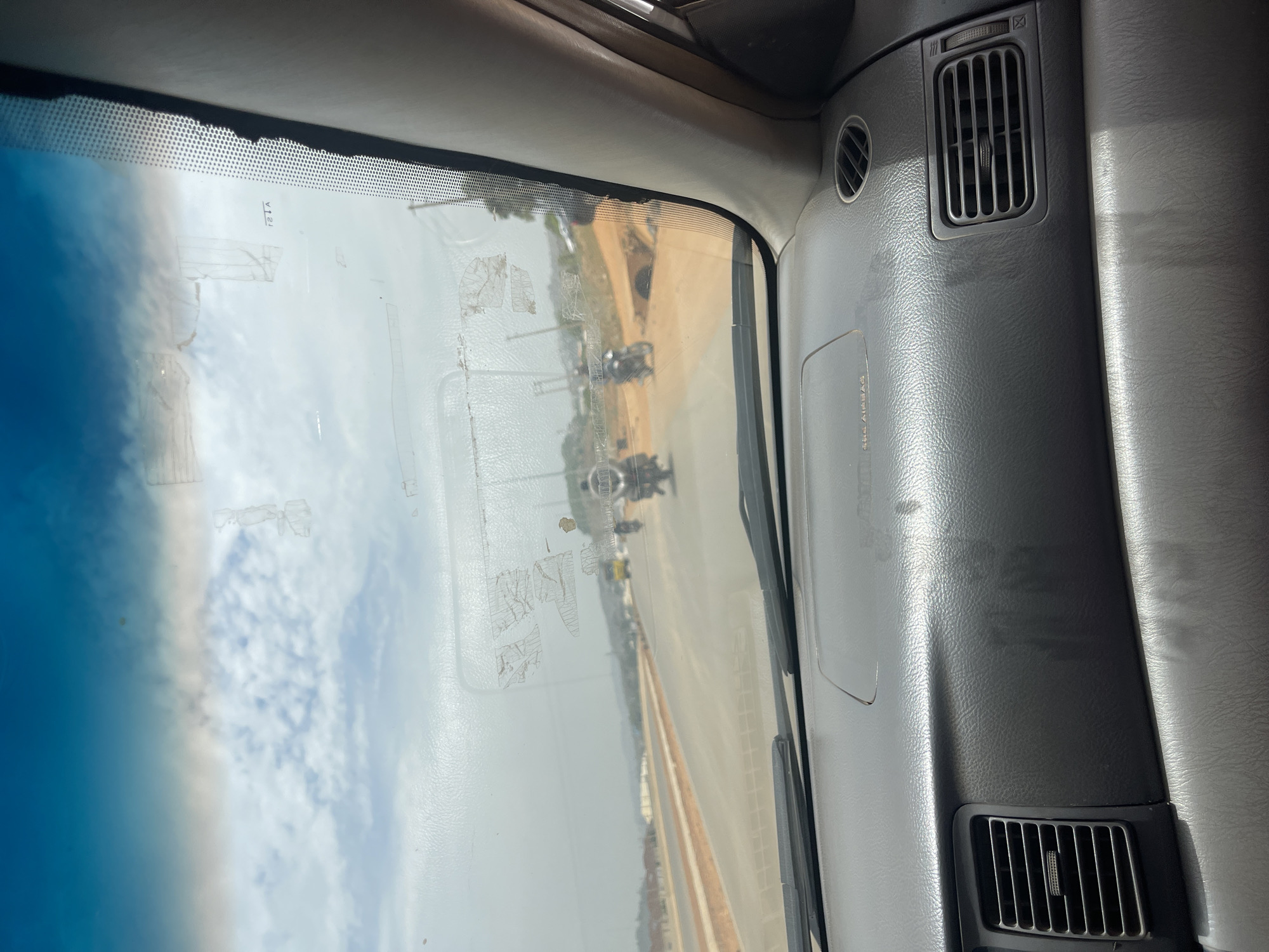 The image presents a view from the interior of a car, looking out through the windshield towards the road and sky. The car's dashboard, predominantly grey, occupies the foreground and right side of the frame, featuring air conditioning vents. A label for "SRS AIRBAG" is visible on the passenger side of the dashboard.
The windshield displays a blue tint at the top, consistent with a sun-blocking band, and has various smudges, reflections, and remnants of adhesive or old stickers. Faint, reversed text, possibly "V12A" or similar, is visible on the upper part of the windshield.
Outside the vehicle, a bright daytime sky with scattered clouds is visible. Below the sky, a road extends into the distance, flanked by unpaved, sandy or dusty terrain. Several vehicles, appearing to be motorcycles or small utility vehicles, are faintly discernible on the road in the mid-distance. There are also indistinct structures or power poles in the background. The lighting suggests it is daytime, possibly late morning or early afternoon, with clear but cloudy weather. The scene depicts a typical road environment from a moving or stationary vehicle.