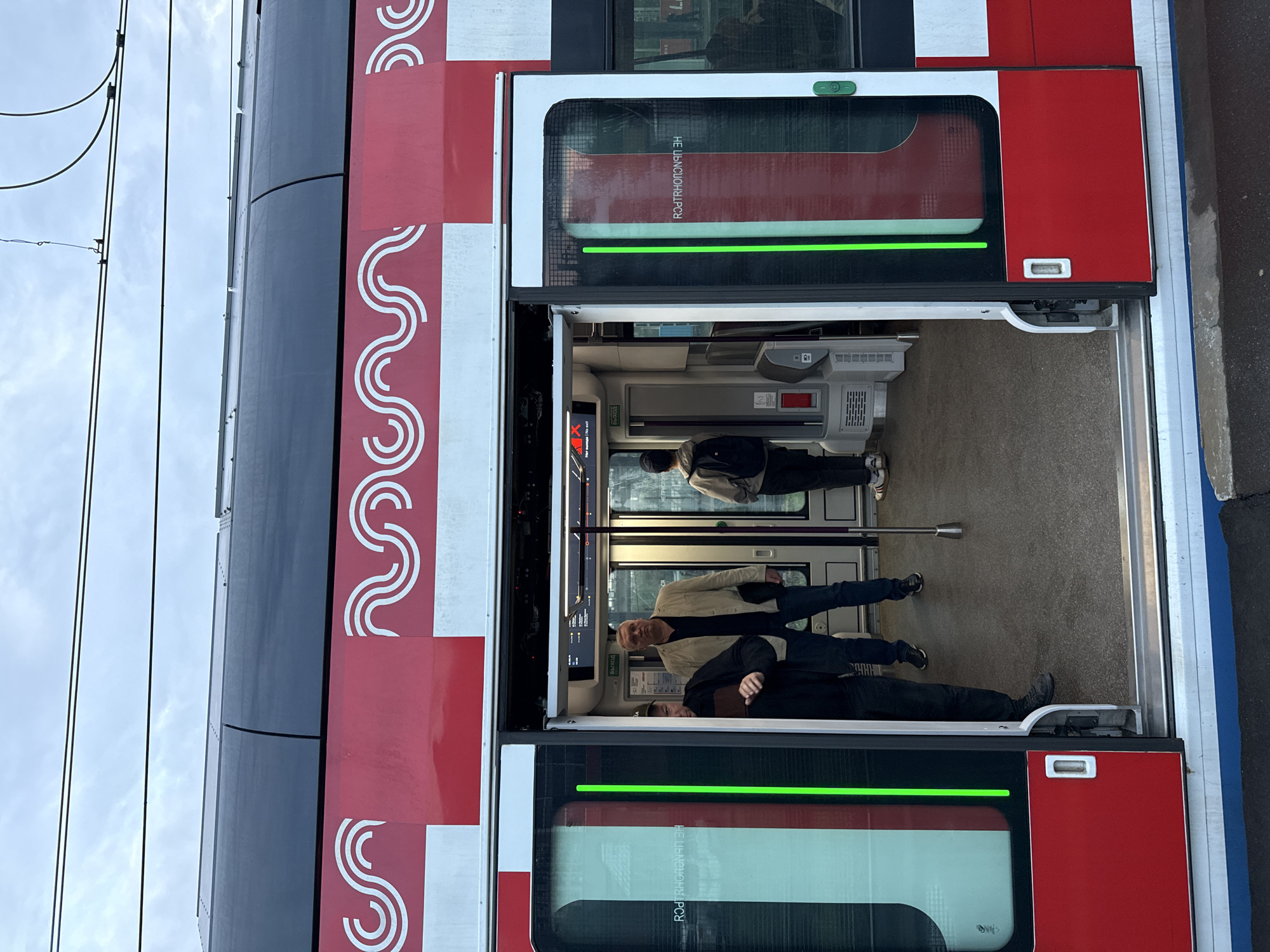 The image shows the open doors of a Moscow Central Diameters (MCD) train. Passengers are visible inside the train. The train's exterior is marked with red and white stripes with a distinctive wavy pattern. Overhead electrical lines are visible above the train. The location is Moscow, Russia, based on the branding of the train and architectural clues.