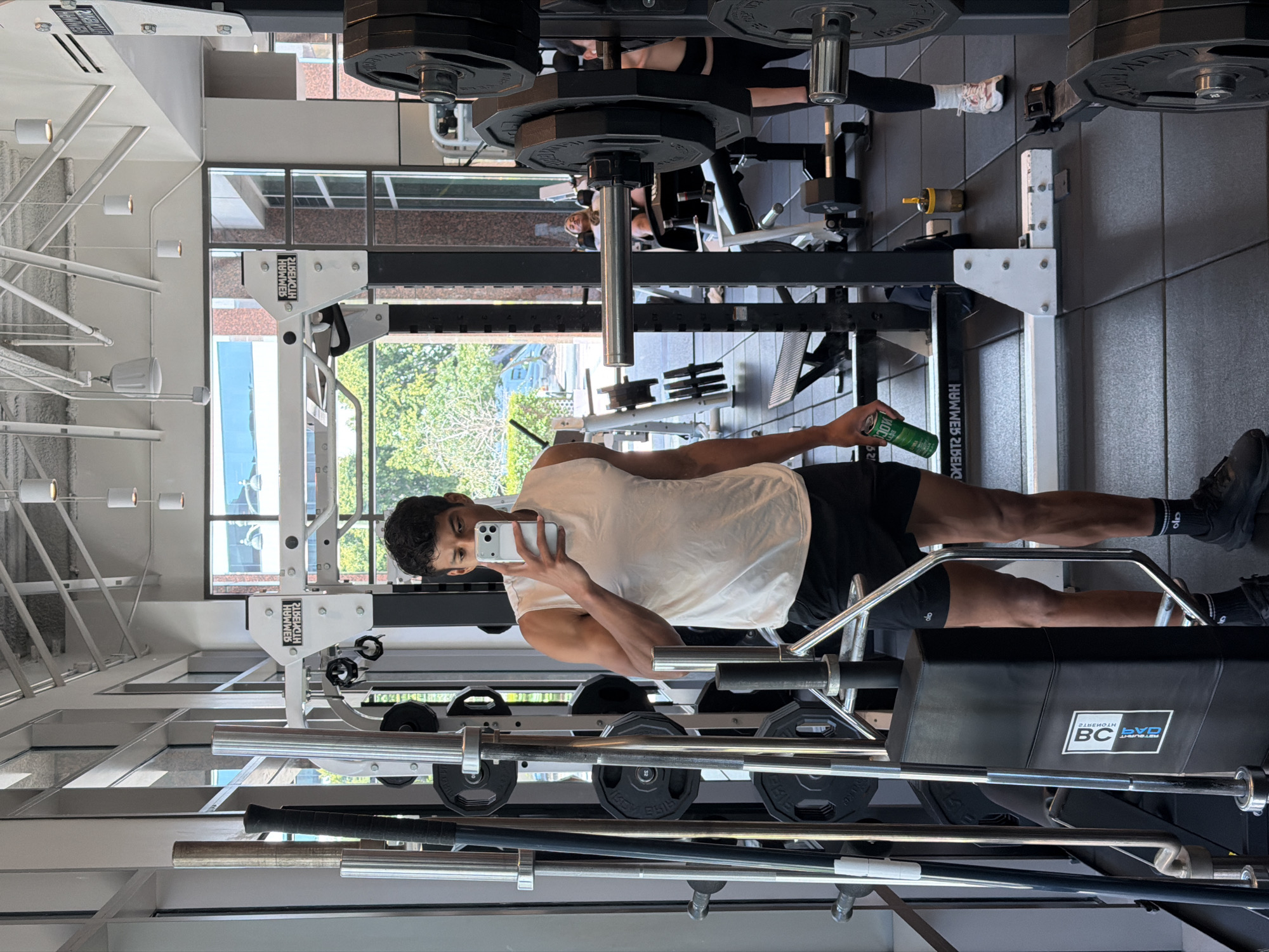 The image shows a man in a gym, holding a phone in his right hand and a green can in his left. He is wearing a white tank top, black shorts, and black shoes with socks. The gym has weightlifting equipment including a power rack with weights, barbells, and plates. A window provides a view of trees and a building exterior. Another person is seen in the background. The gym's location is in Los Angeles, United States of America, based on the building exterior and gym equipment.