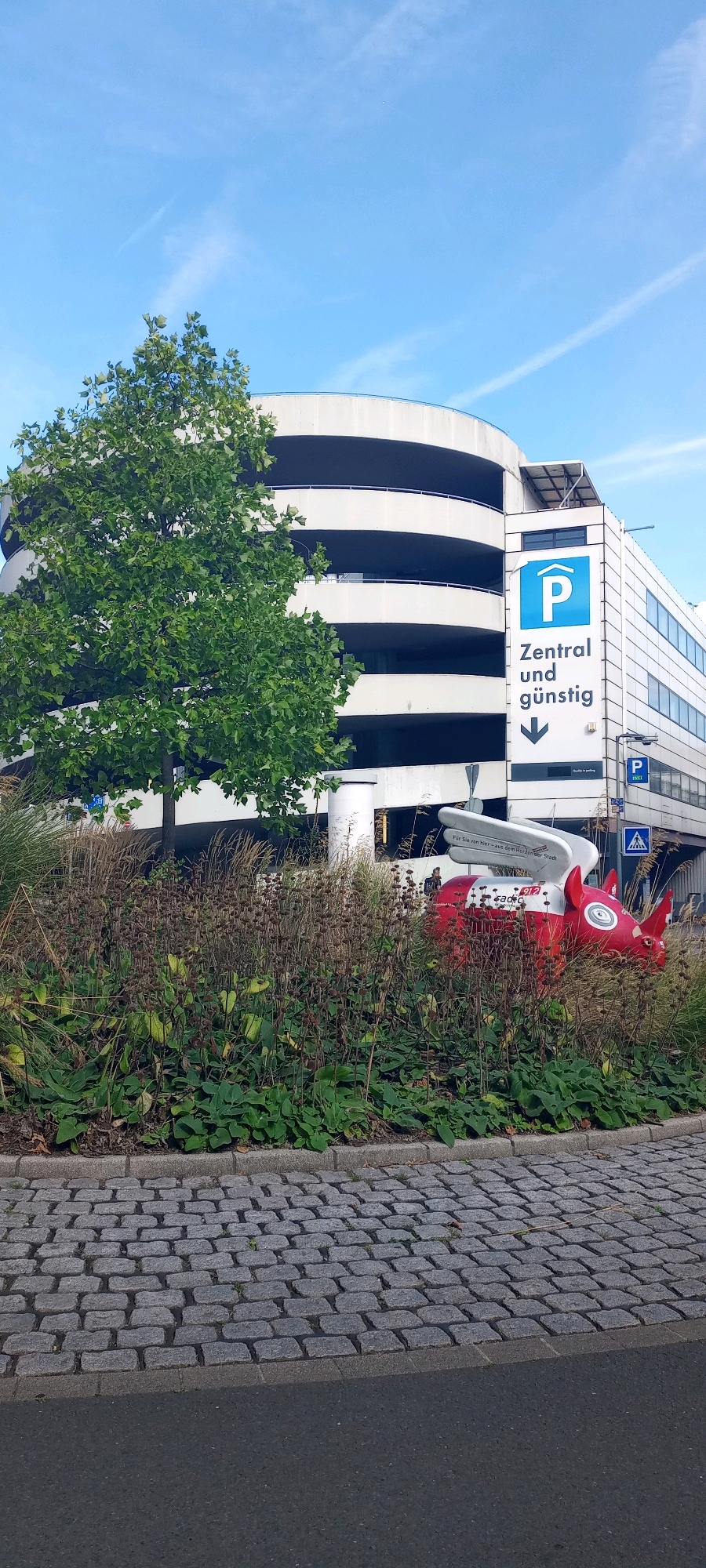 The image shows a multi-story concrete parking garage with a curved facade on the left and a rectangular section on the right. A large green tree is positioned in front of the garage on the left side. On the right side of the garage, a prominent sign with a blue parking symbol and the text "Zentral und günstig" (Central and affordable) is displayed. Below this sign, an arrow points downwards. Further down the facade, another smaller parking sign is visible.
In the foreground, a dense planting of ornamental grasses and green foliage lines the base of the garage. Peeking out from behind the foliage on the right is a brightly colored red and white sculpture of a stylized rhinoceros with wings, suggesting it's a decorative element or art installation. There are hints of people in the background of the garage, and a small pedestrian crossing sign is visible on the right side of the building. The ground in front of the vegetation is paved with grey cobblestones, transitioning to asphalt on the far right. The sky above is blue with wispy white clouds.
The location is the parking garage at Dortmund Airport.