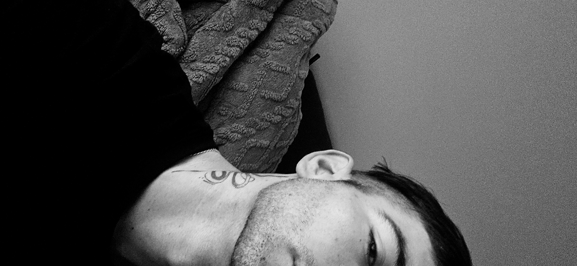 This black and white, close-up photograph captures a person, likely male, lying down in an indoor setting in Plovdiv, Bulgaria. The subject's head, neck, and a portion of their left shoulder are visible, indicating a pose of rest or relaxation, possibly on a bed or couch. The person has short, dark hair, potentially styled with shaved sides, and displays light facial stubble. Their left eye is partially visible, appearing to gaze towards the viewer or slightly downwards, conveying a neutral and relaxed expression.
A notable detail is a tattoo positioned on the side of the person's neck, extending onto the upper shoulder. The tattoo features two stylized, somewhat oval shapes that resemble eyes, connected by a horizontal line. Each 'eye' contains a smaller, darker circular or semi-circular element, suggesting pupils or irises. A subtle, light-colored chain or necklace is also visible around the person's neck, resting against their skin.
Behind the person's head and shoulder, a textured blanket or fabric is visible, providing a soft, patterned element to the composition. To the right side of the frame, a plain, light-toned wall or surface serves as the background. The lighting is soft and ambient, characteristic of an indoor environment, and the black and white conversion enhances the textures and tonal contrasts within the image. No specific time of day or external weather conditions are discernible.