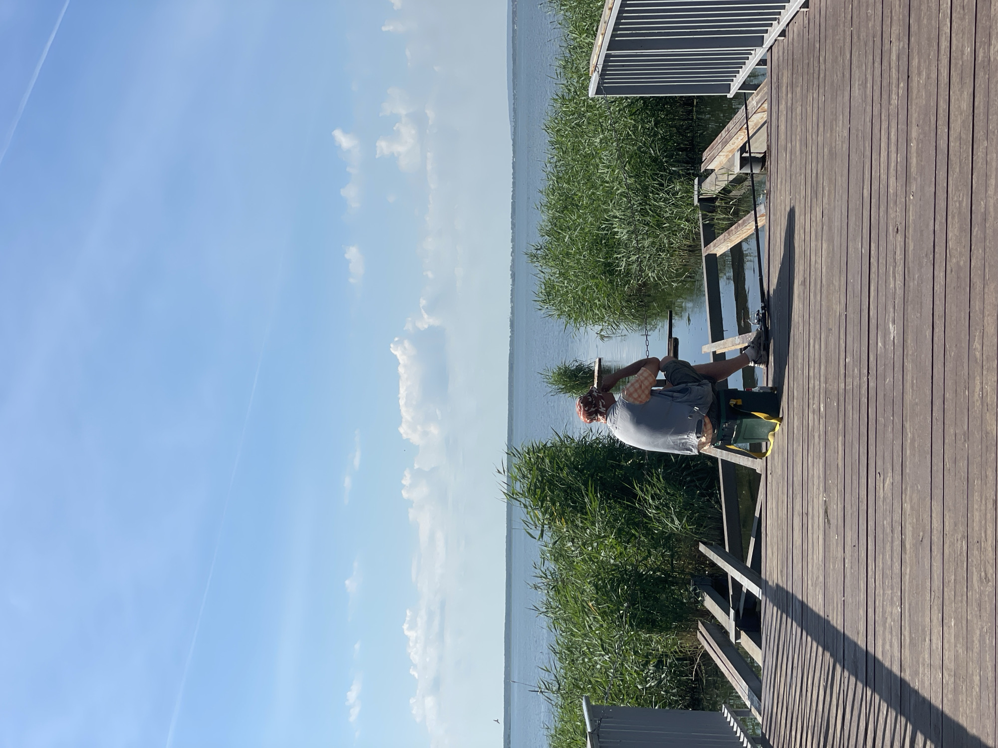 A person sits on a wooden dock facing a body of water. The person is wearing a grey vest over a checkered shirt, shorts, and a patterned hat. They are seated on a green tackle box. A fishing rod extends from the dock into the water. Tall green reeds line the edge of the water, and a distant shoreline is visible under a blue sky with scattered white clouds. The dock itself is constructed of wooden planks and has metal railings on the right side. Some sections of the dock appear unfinished or damaged, with exposed metal supports.