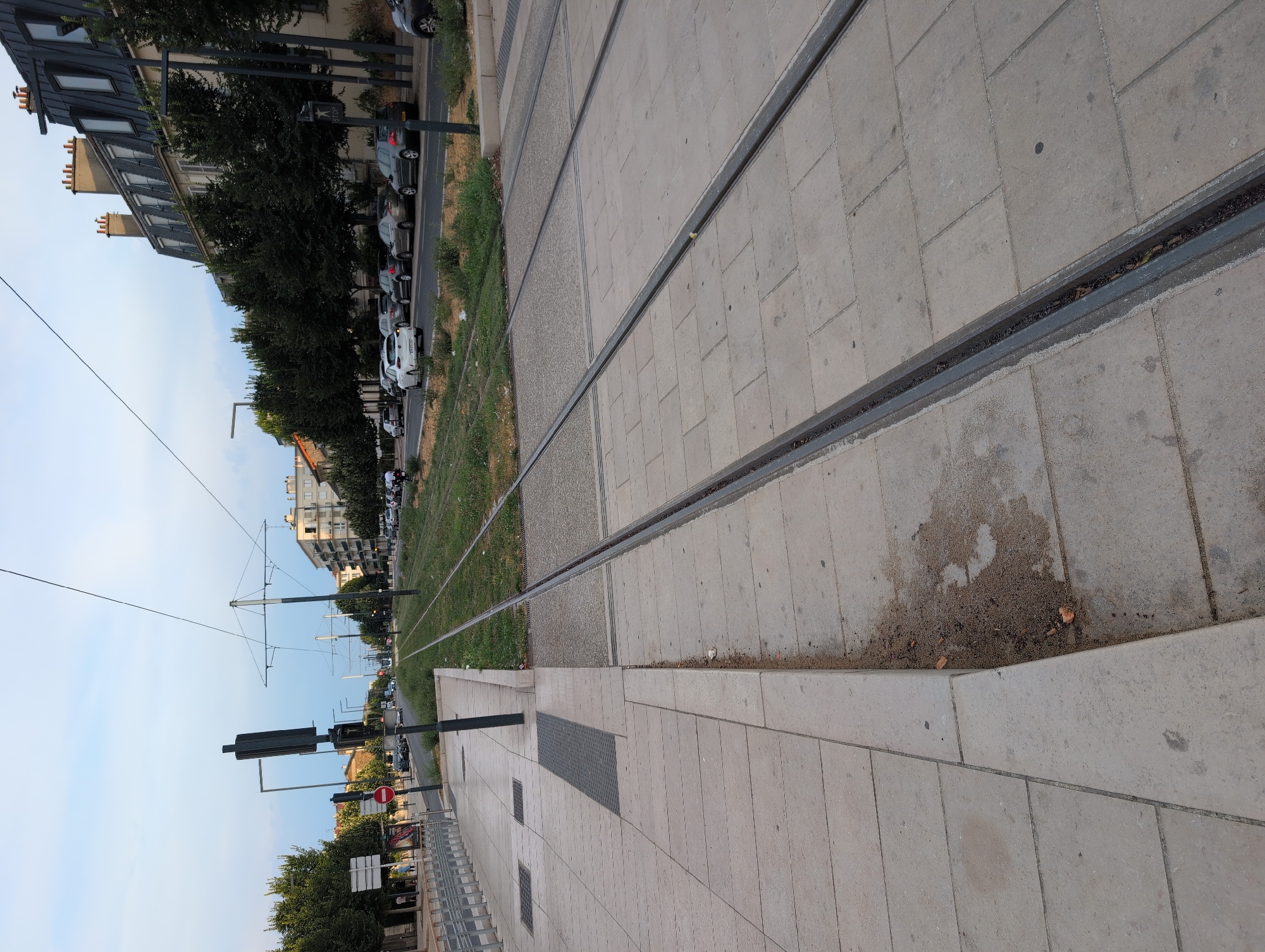 This image depicts a street scene in Choisy-le-Roi, France, featuring tram tracks running down the center of a road. On the left side of the image is a paved sidewalk with bollards and a bus stop shelter. A traffic signal with a red stop sign is visible. The tram tracks are flanked by grass and concrete. On the right side of the road, there are parked cars and a building with a mansard roof. The sky is overcast, and trees line the street. No individuals are prominently featured in the image.